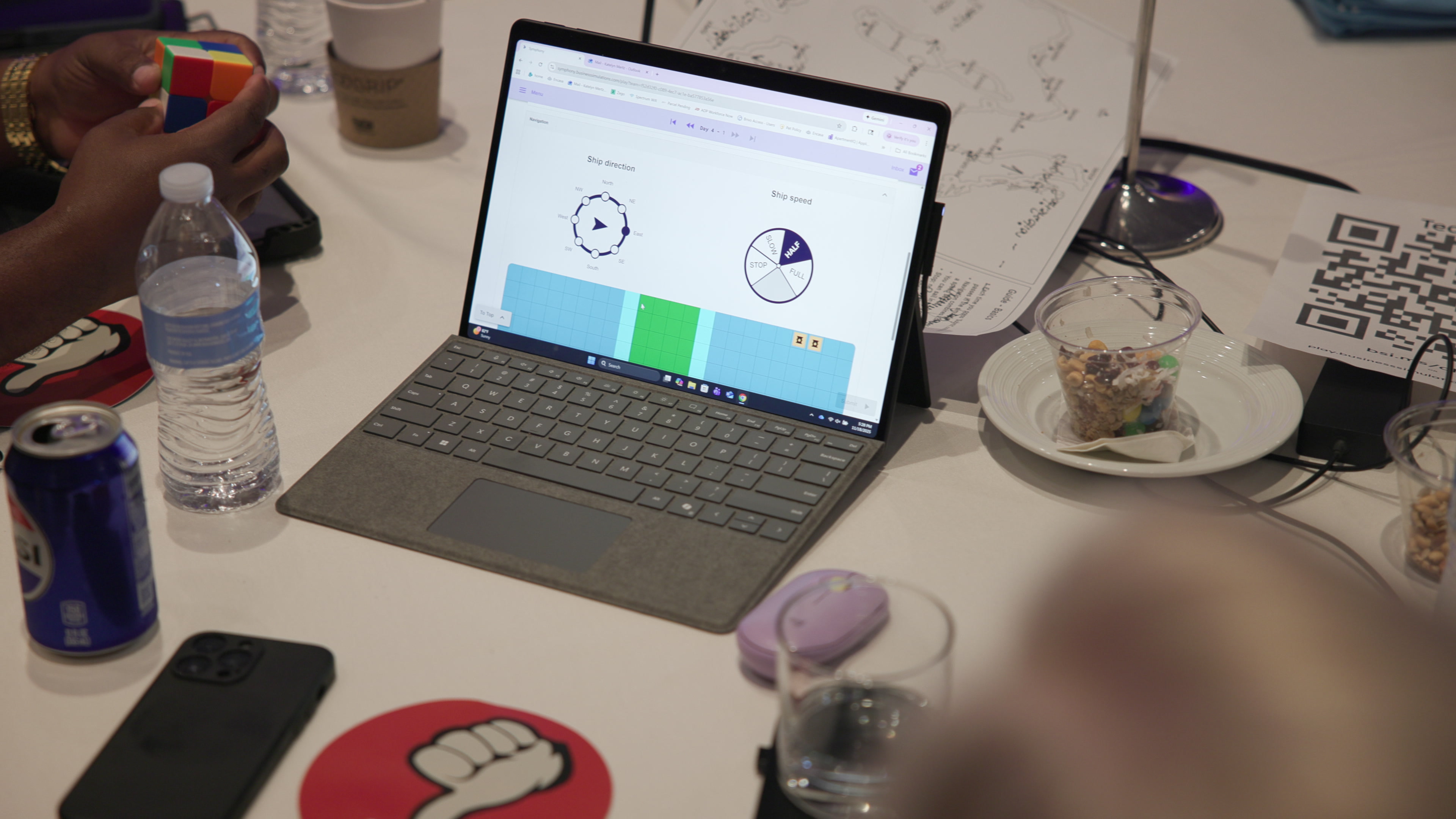 A close-up view of a table showing a laptop with a ship navigation dashboard, alongside drinks, snacks, and a person's hands holding a Rubik's cube.