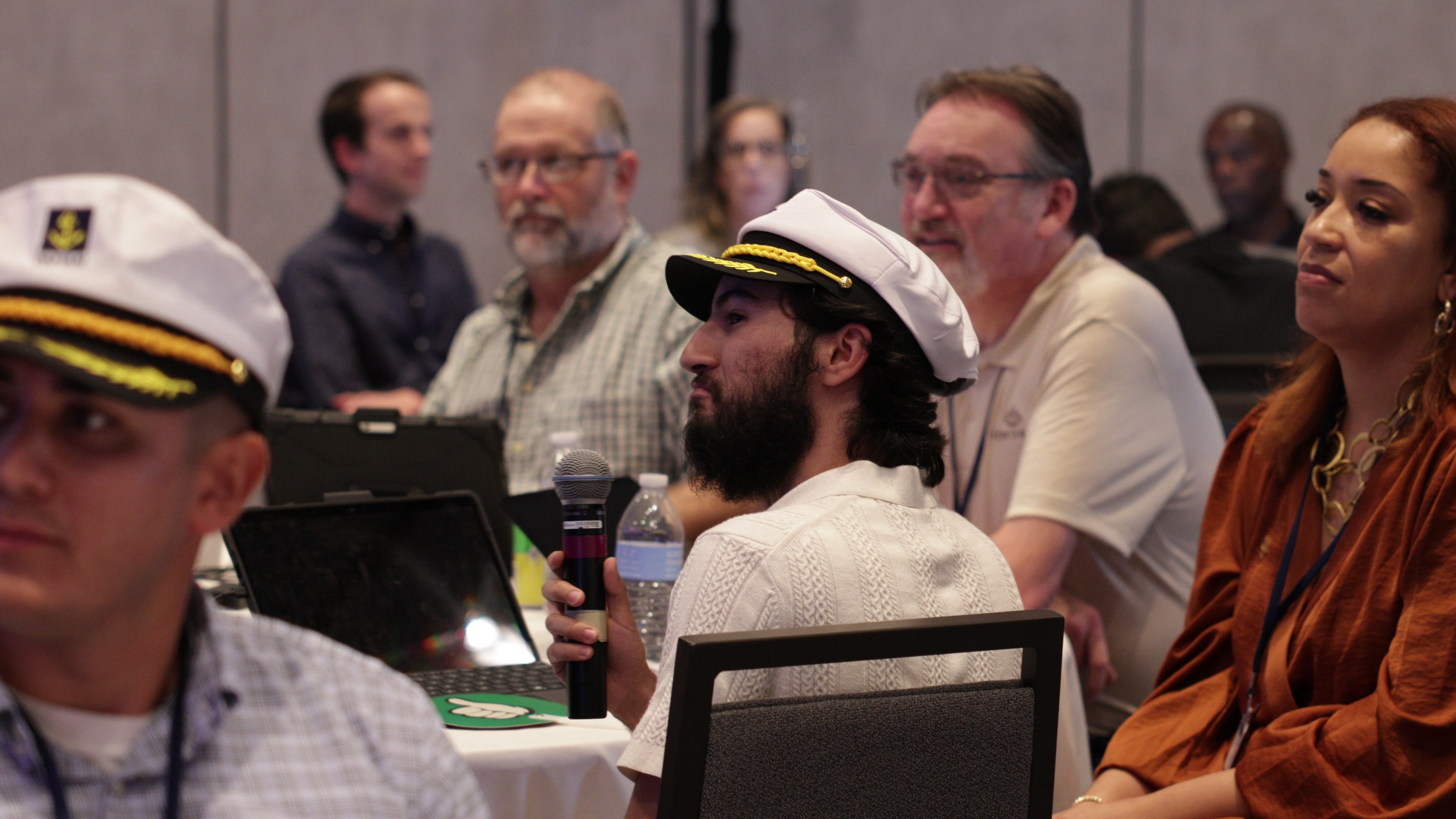 A man wearing a captain's hat holds a microphone at a table, with other attendees sitting nearby.