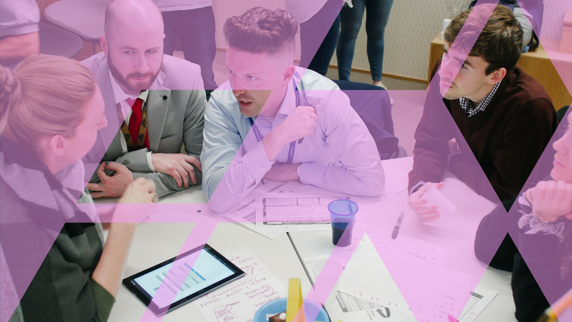 Four business simulation participants leaning into an intense discussion across a table of documents and a tablet, overlaid with a pink Business Simulations brand motif
