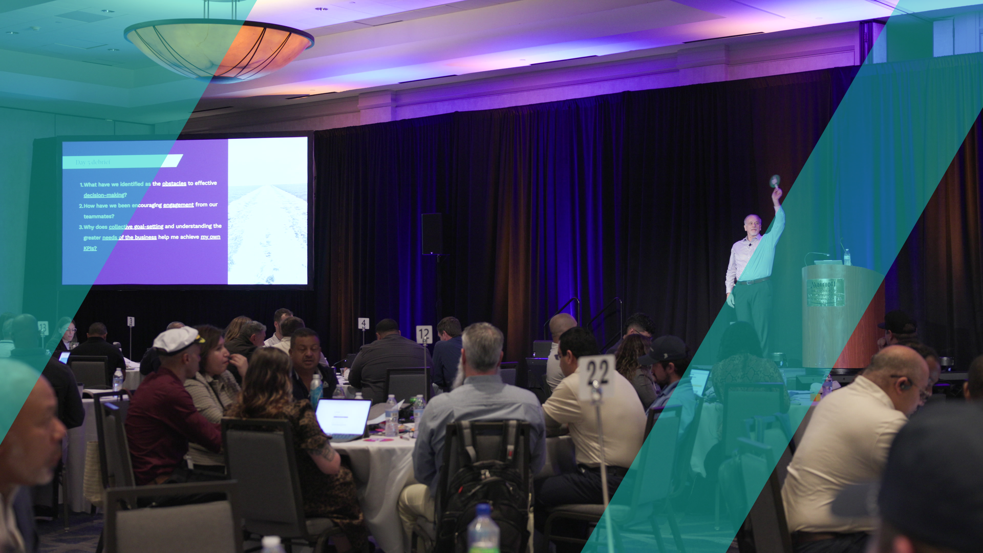 A facilitator presenting to a large ballroom of participants seated at round tables during a business simulation.