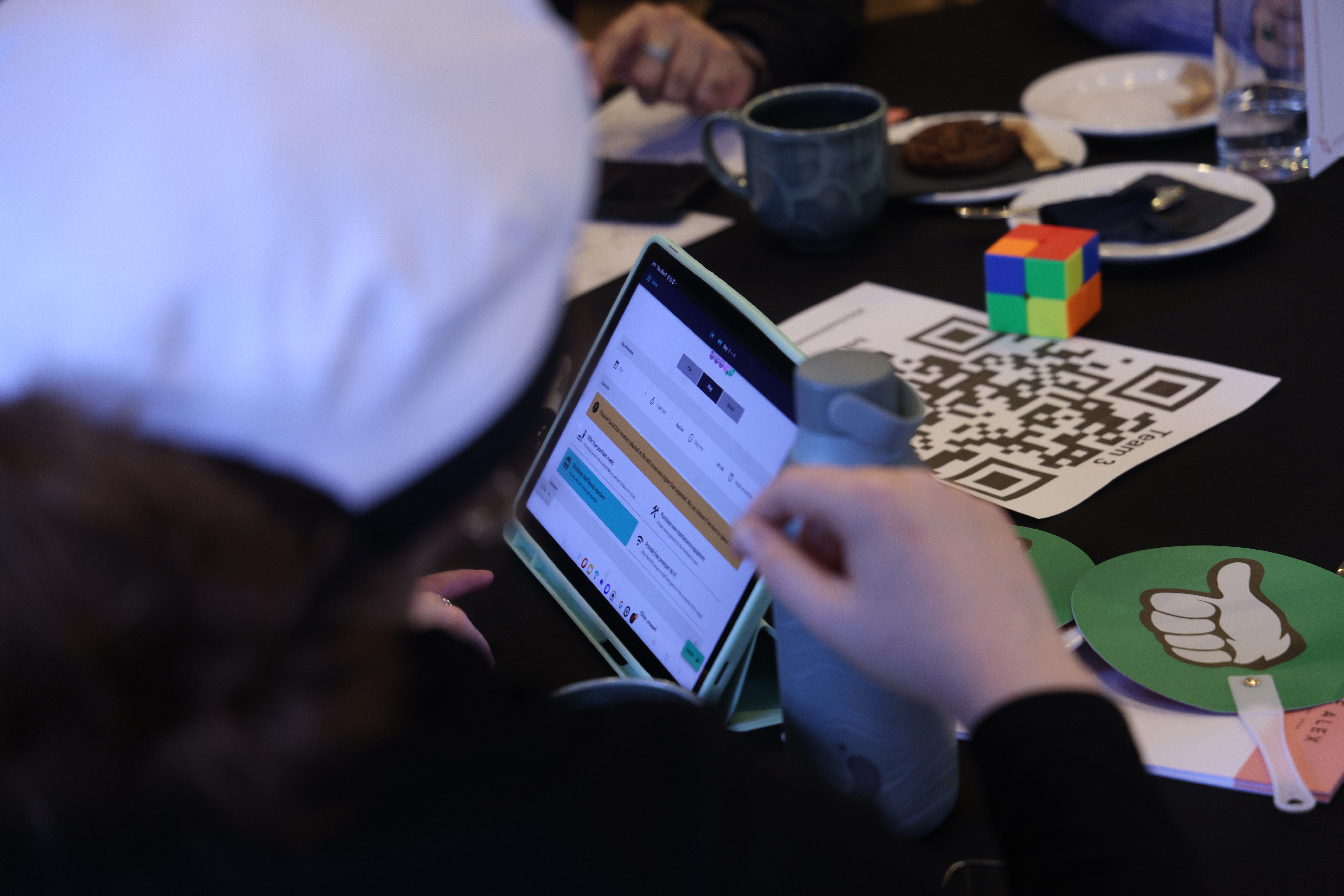Participant views business simulation dashboard on tablet during team exercise at table with materials and refreshments.