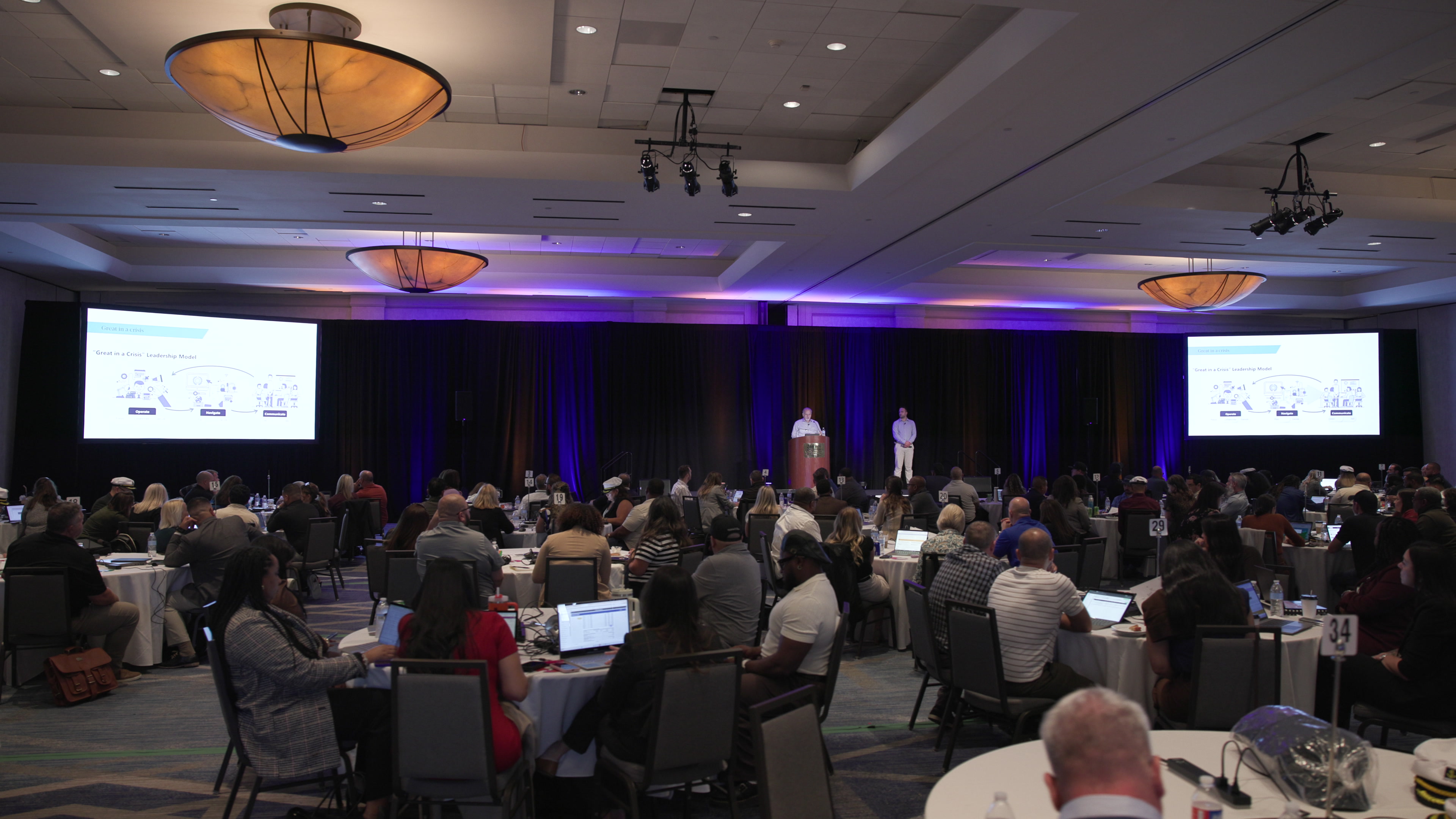 A wide view from the back of a large conference ballroom, showing attendees seated at round tables facing a stage with a presenter at a podium and two large projector screens.
