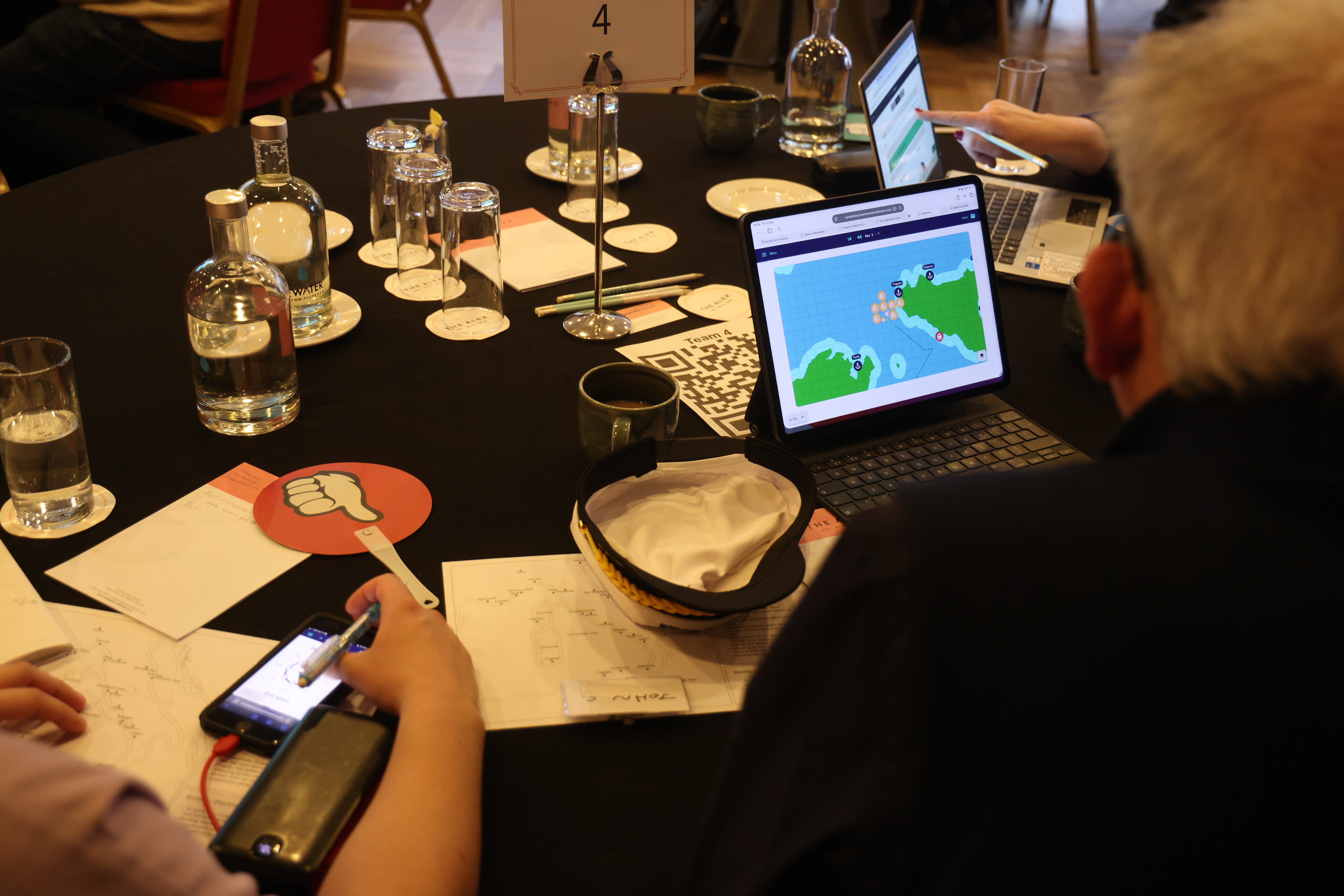 Team members at a black table during a business simulation, viewing strategy maps on laptops while taking notes.