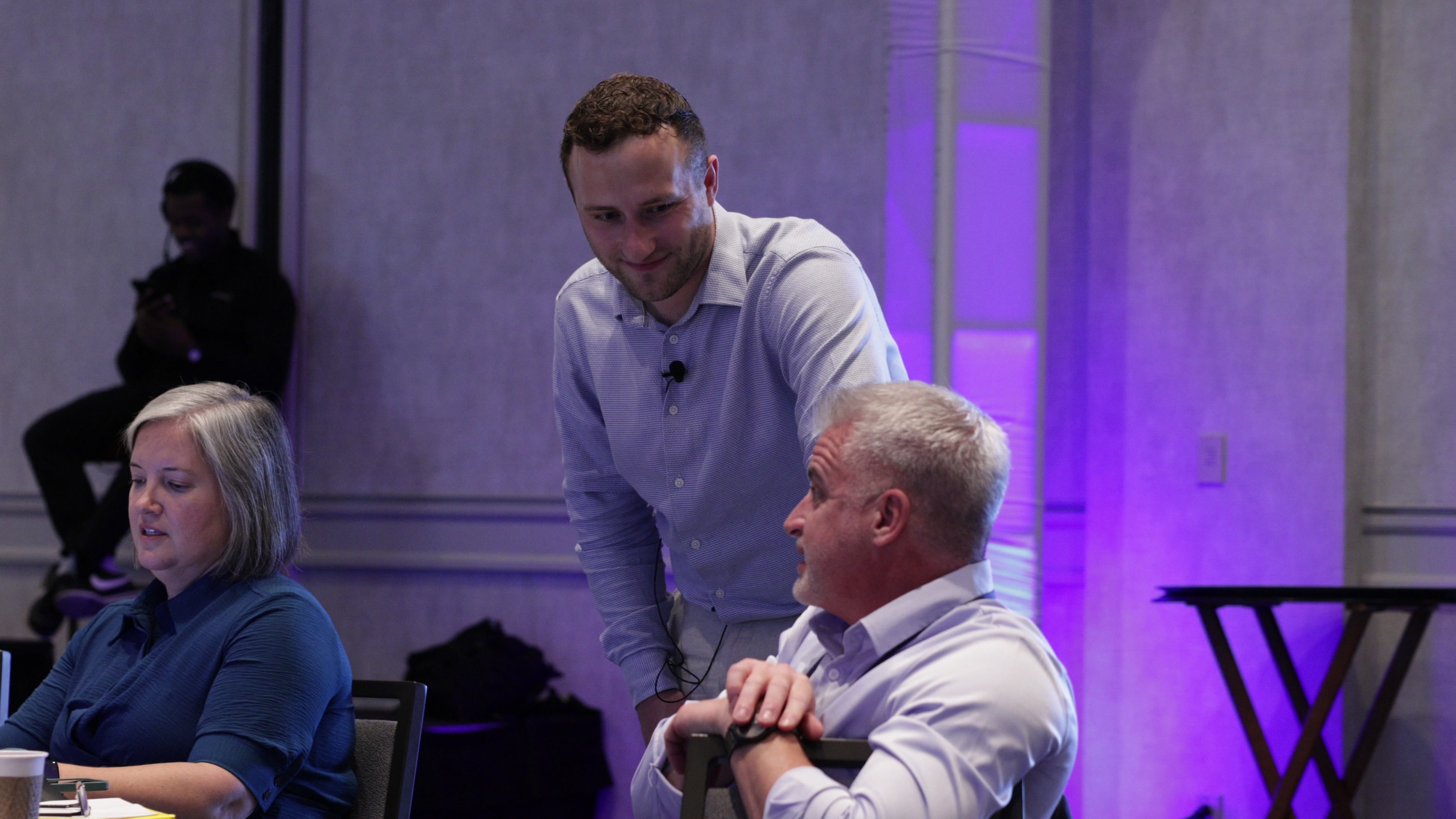 A facilitator leans in to speak with a seated participant at a round table in a conference room.