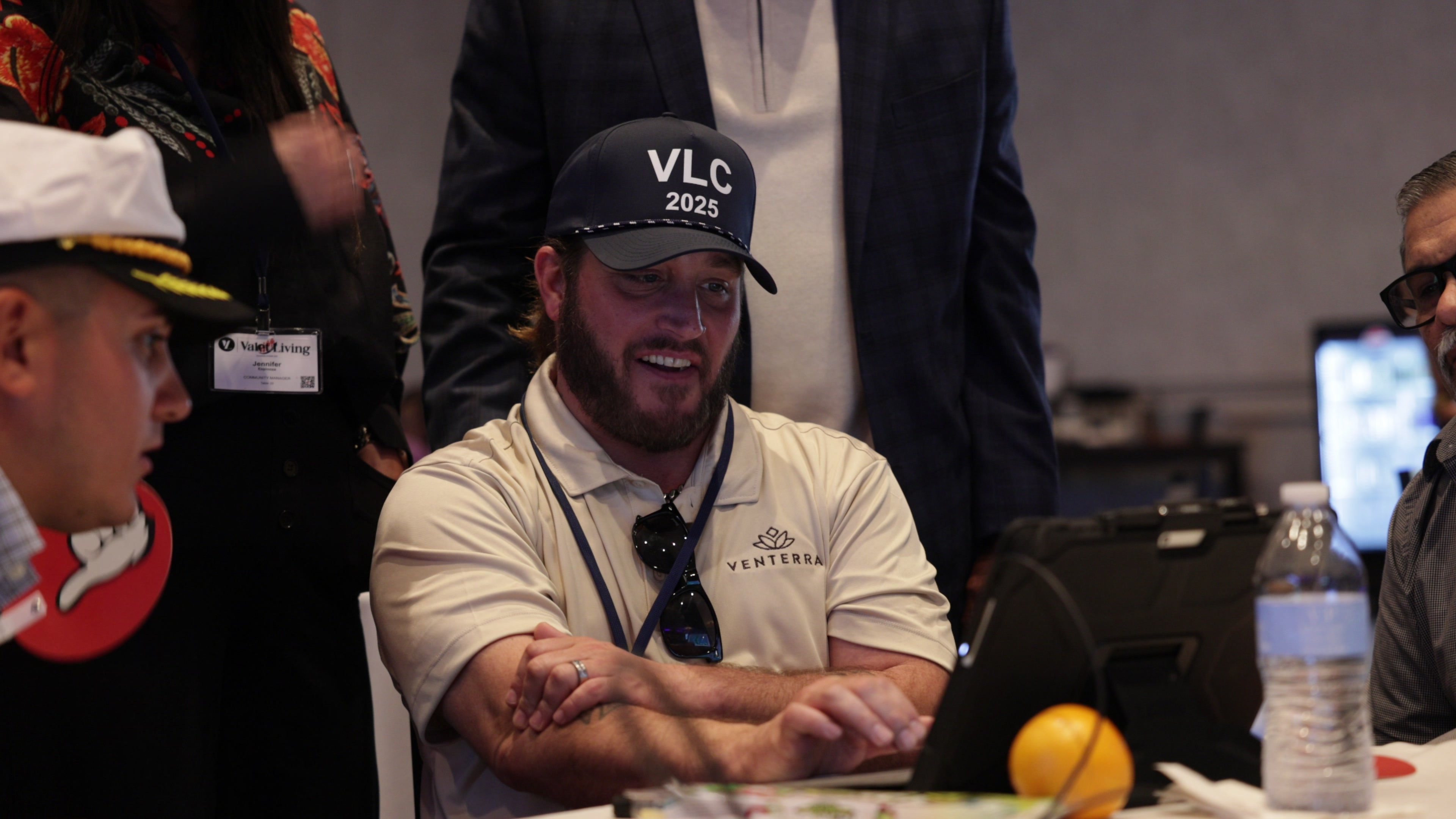 A man in a branded cap and polo shirt smiles while using a tablet at a round table with other participants.