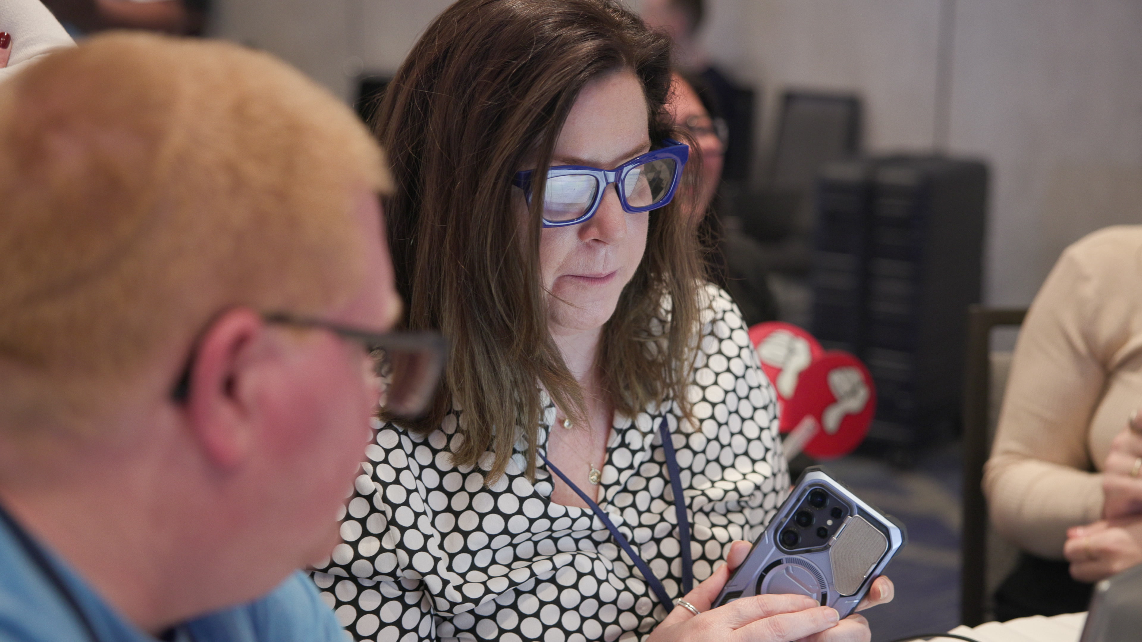 A woman wearing blue glasses looks thoughtfully at a smartphone she is holding, viewed over the shoulder of a man in the foreground.
