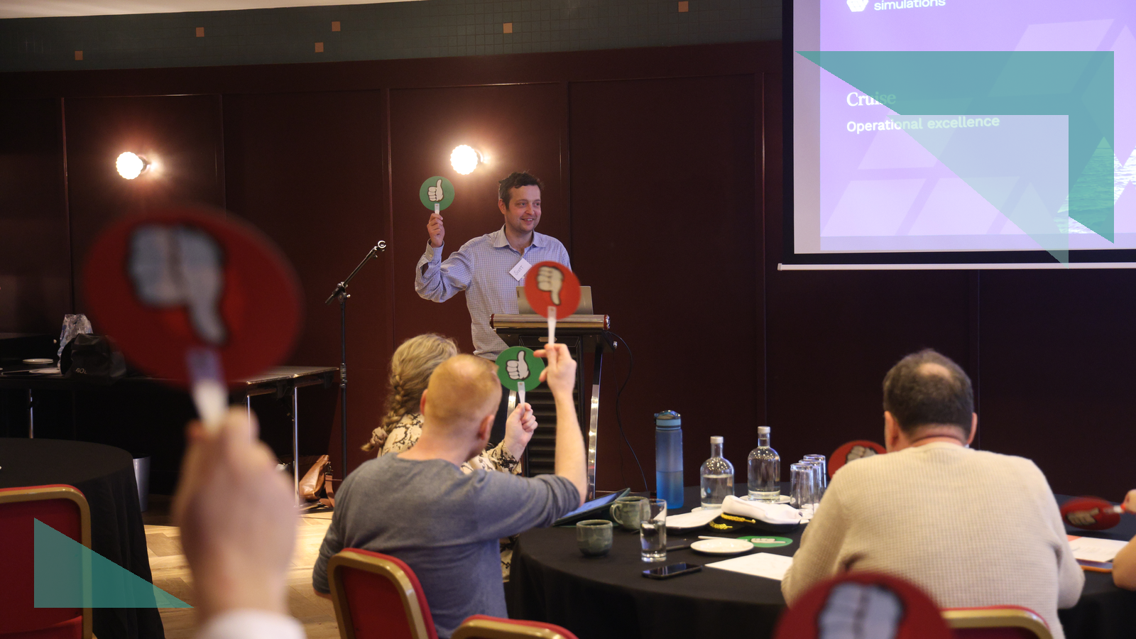A facilitator holds up a thumbs-up card as participants vote during a live business simulation, with the Business Simulations brand motif overlaid in teal