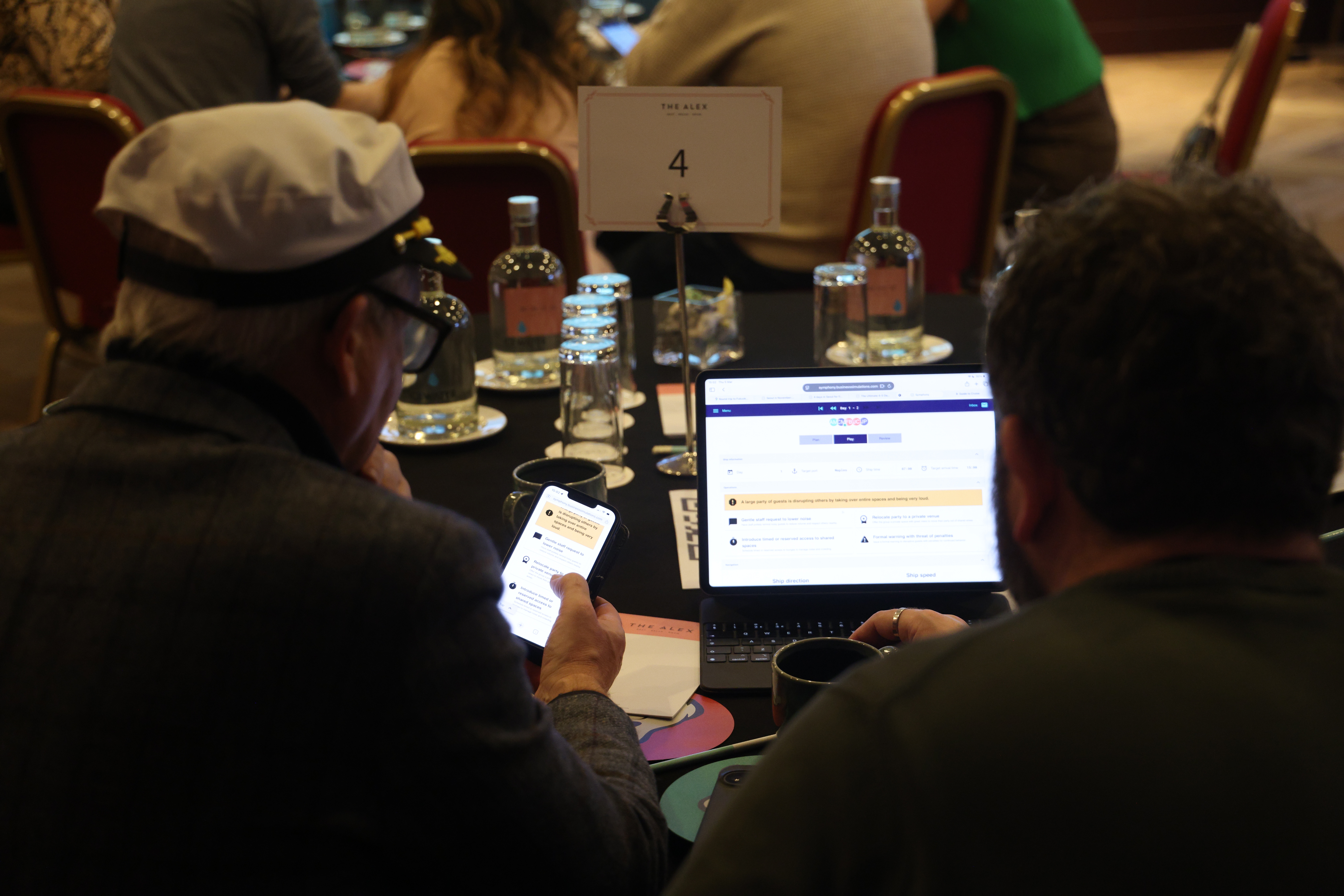 Team members collaborate at a business simulation using laptops and mobile devices during a facilitated workshop session.