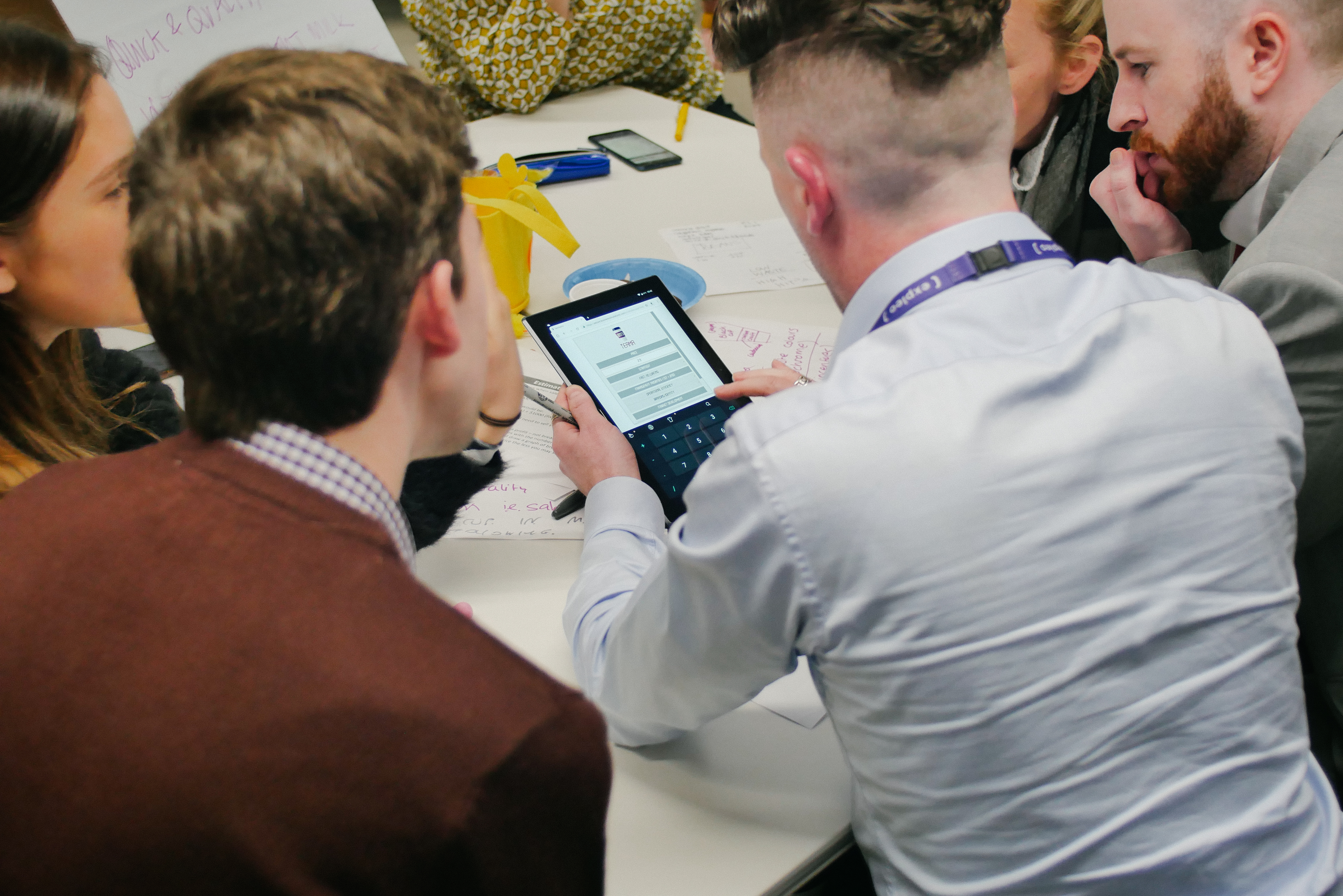 Team of five professionals huddle around a tablet device reviewing business simulation results during a facilitated workshop.