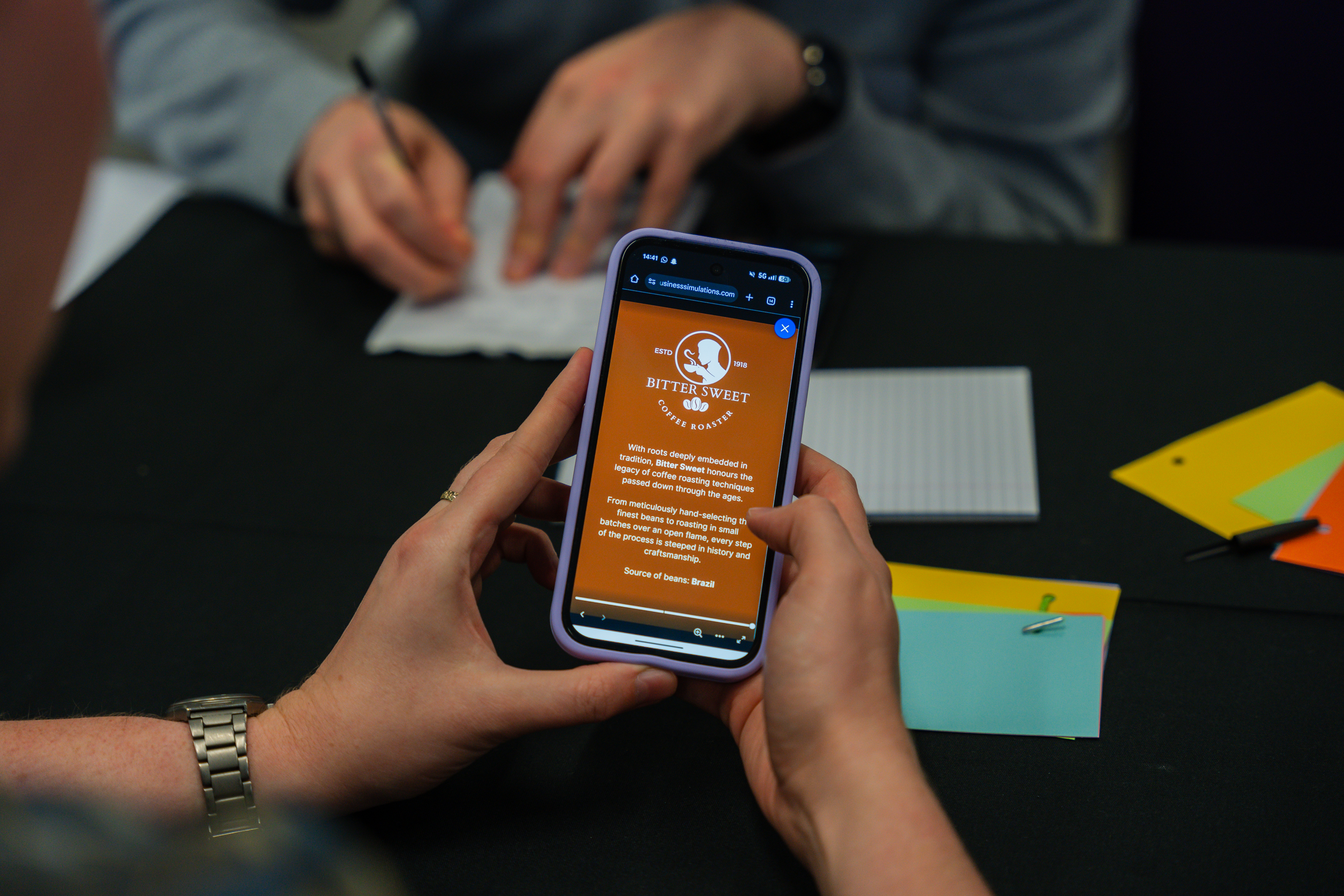 Team members view a mobile app displaying a business simulation interface during a collaborative work session.