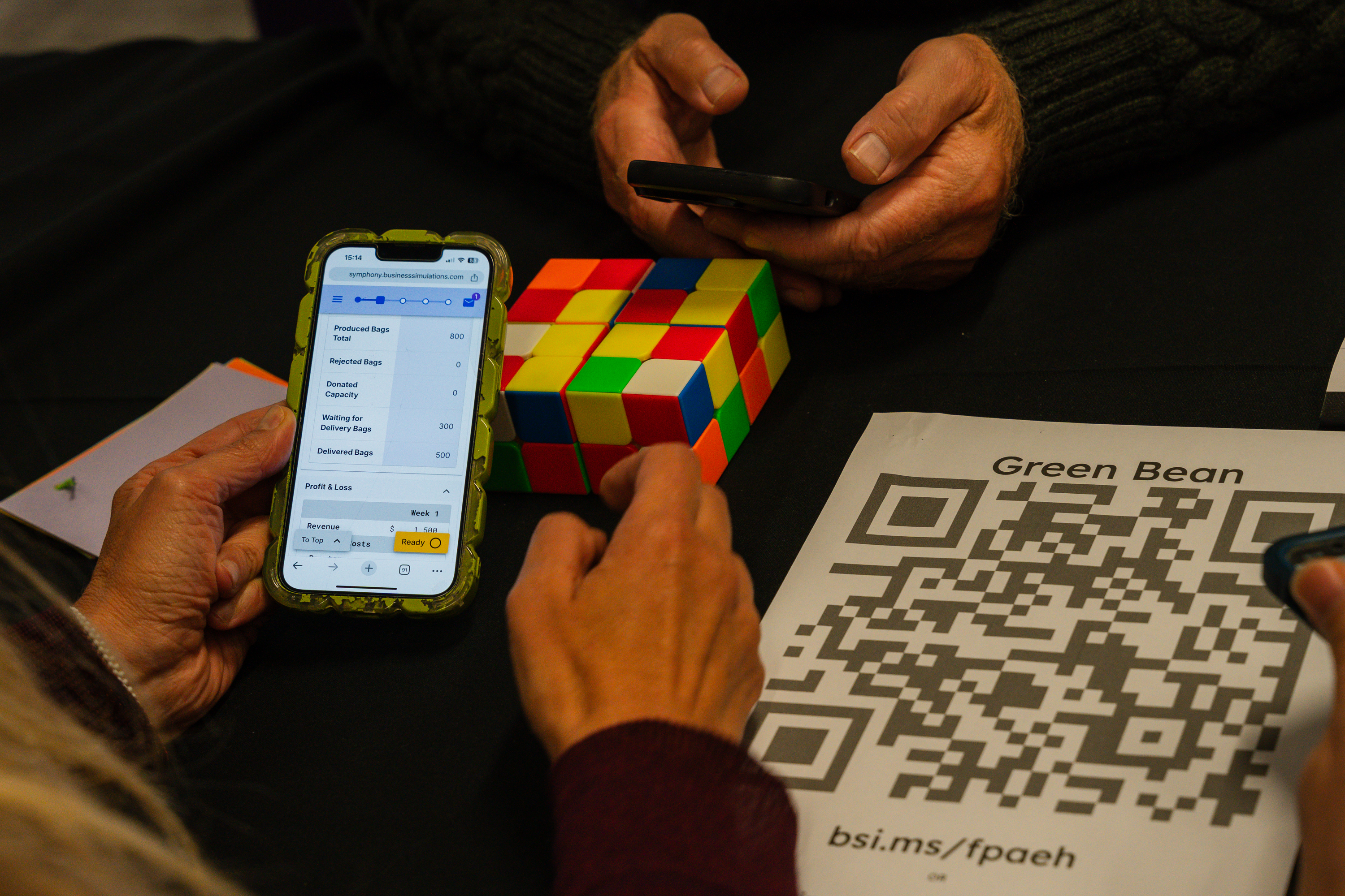 Participant holding smartphone displaying simulation interface while solving a Rubik's cube during a business simulation activity.