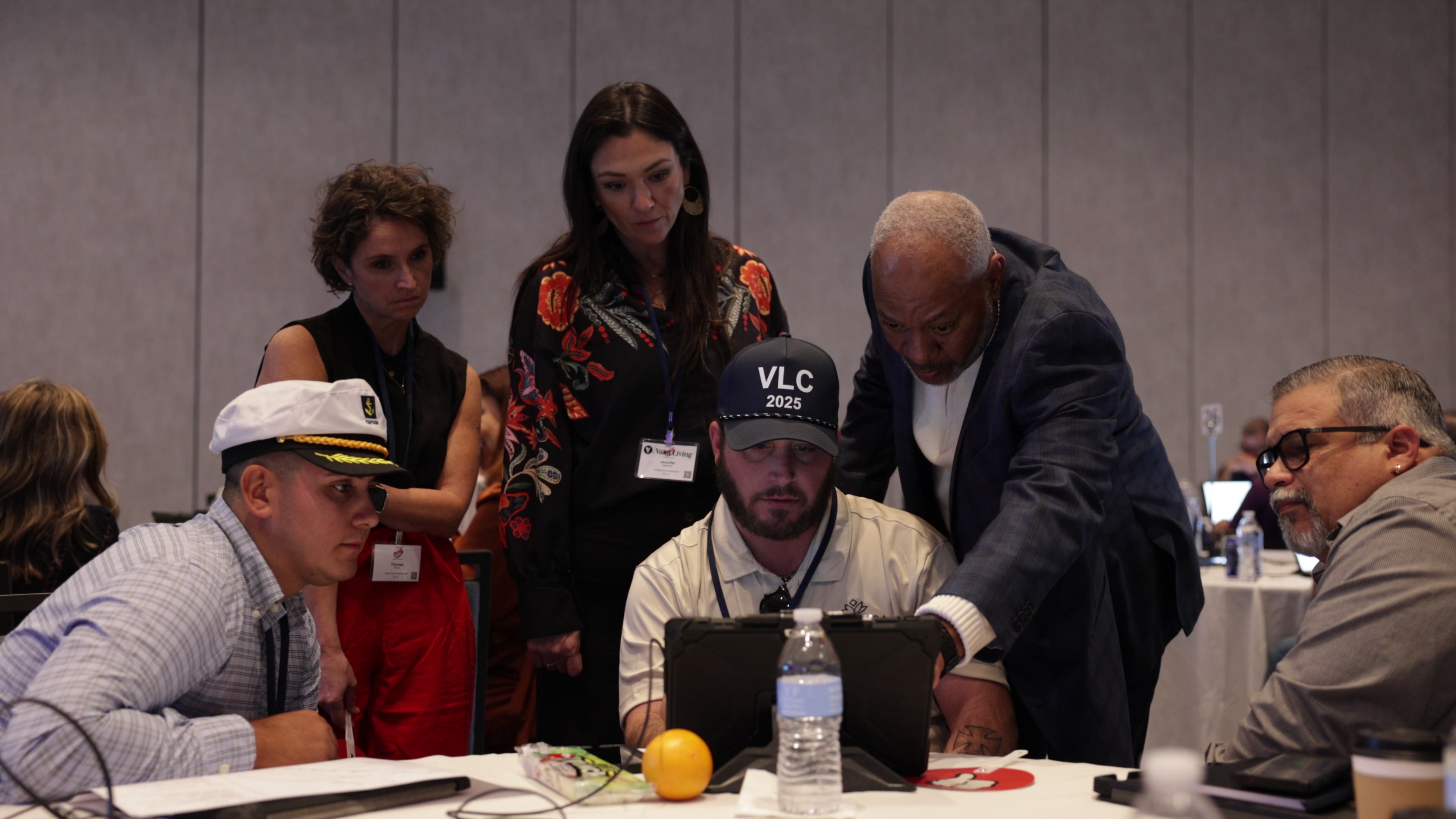 Five people gather around a tablet on a round table, with one man standing and pointing at the screen.