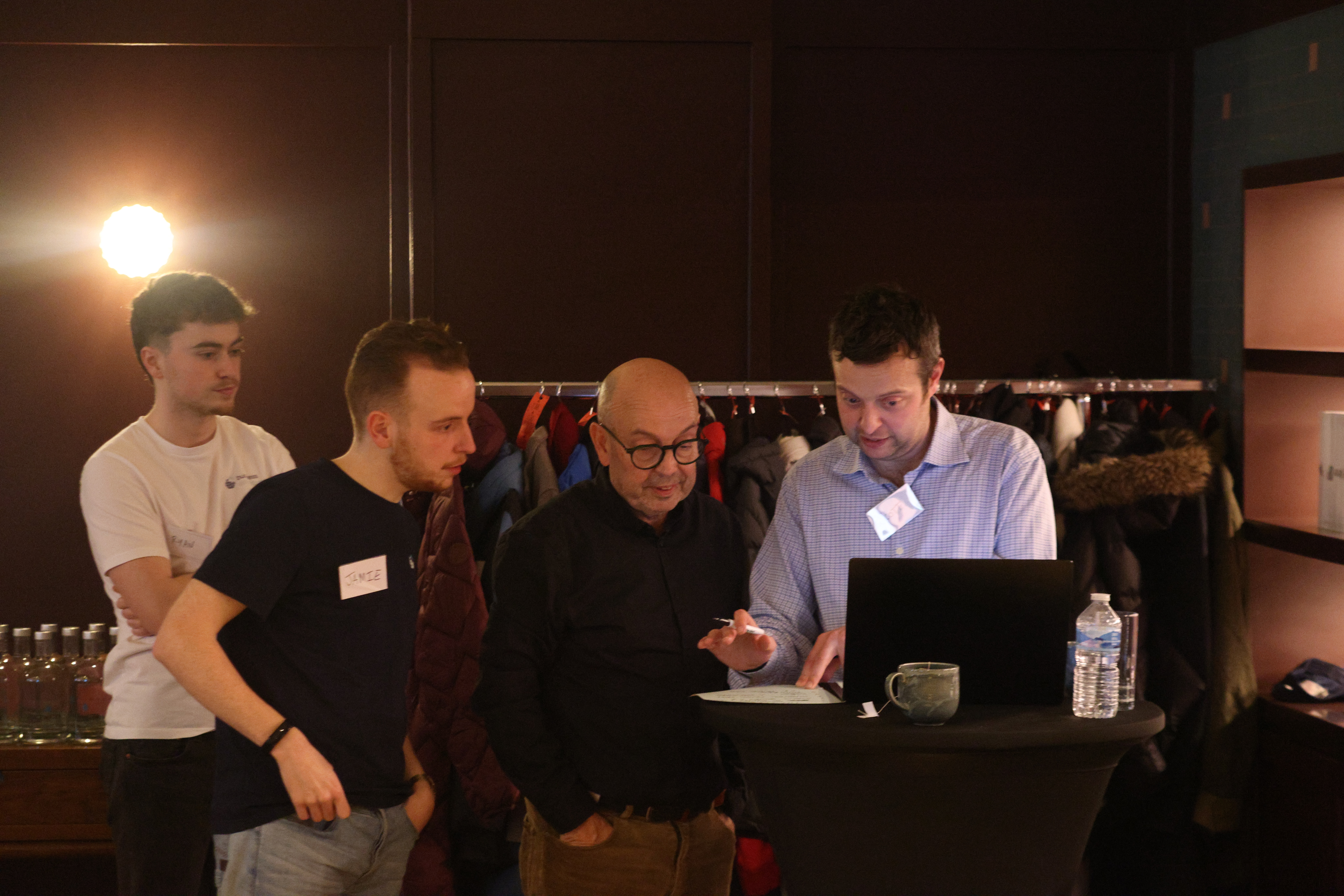 Four men gathered around a laptop during a business simulation event, with participants visible in the background.