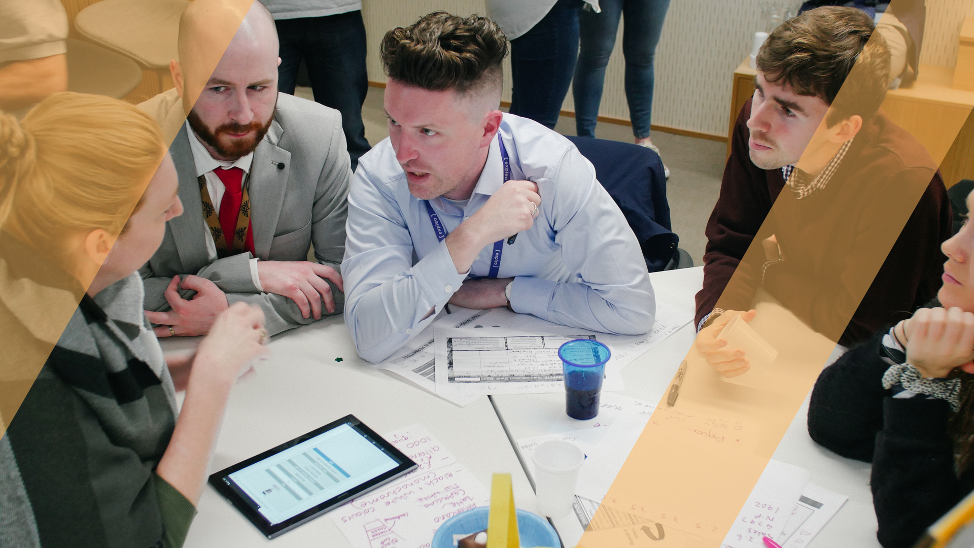 A team leaning in around a table to work through a problem together, with a tablet and working papers, under an amber brand-motif overlay