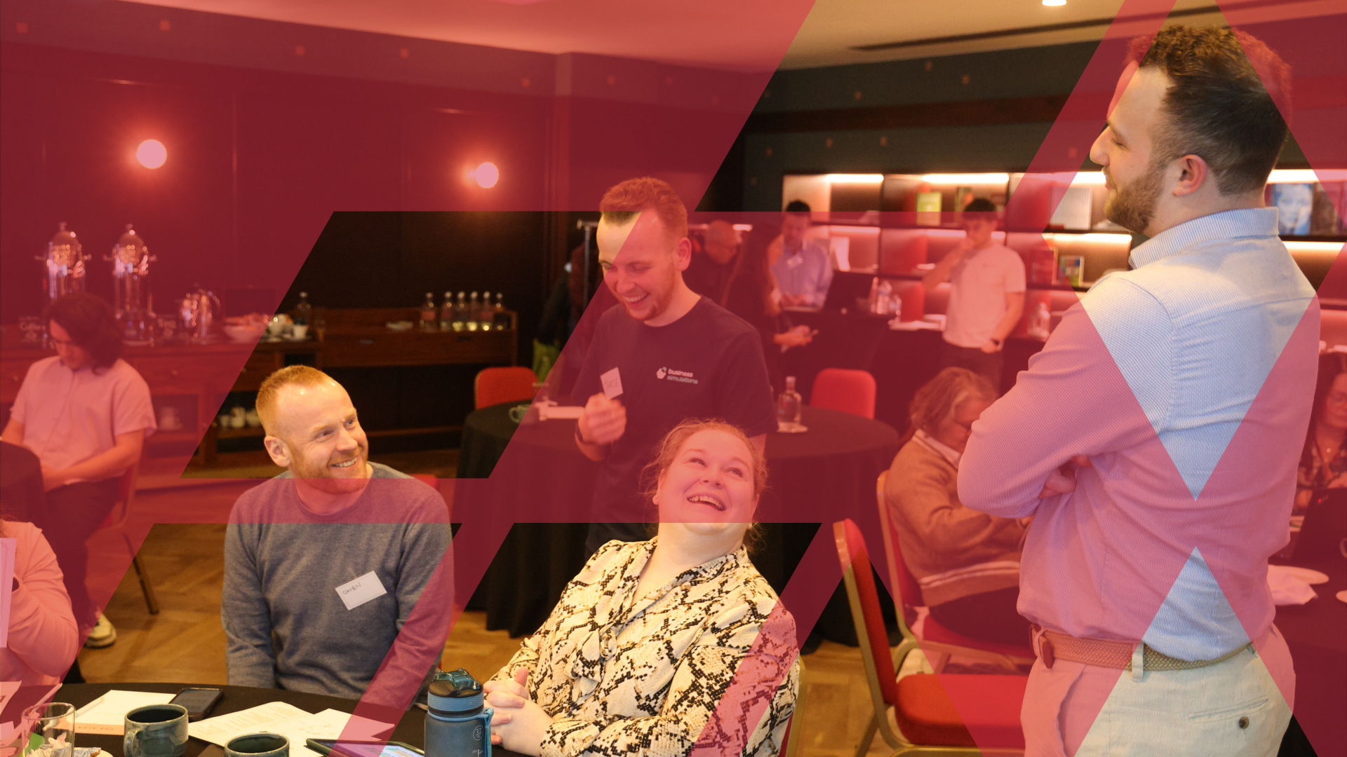 A team leader engaging a group of smiling colleagues during a live business simulation, overlaid with a red Business Simulations brand motif