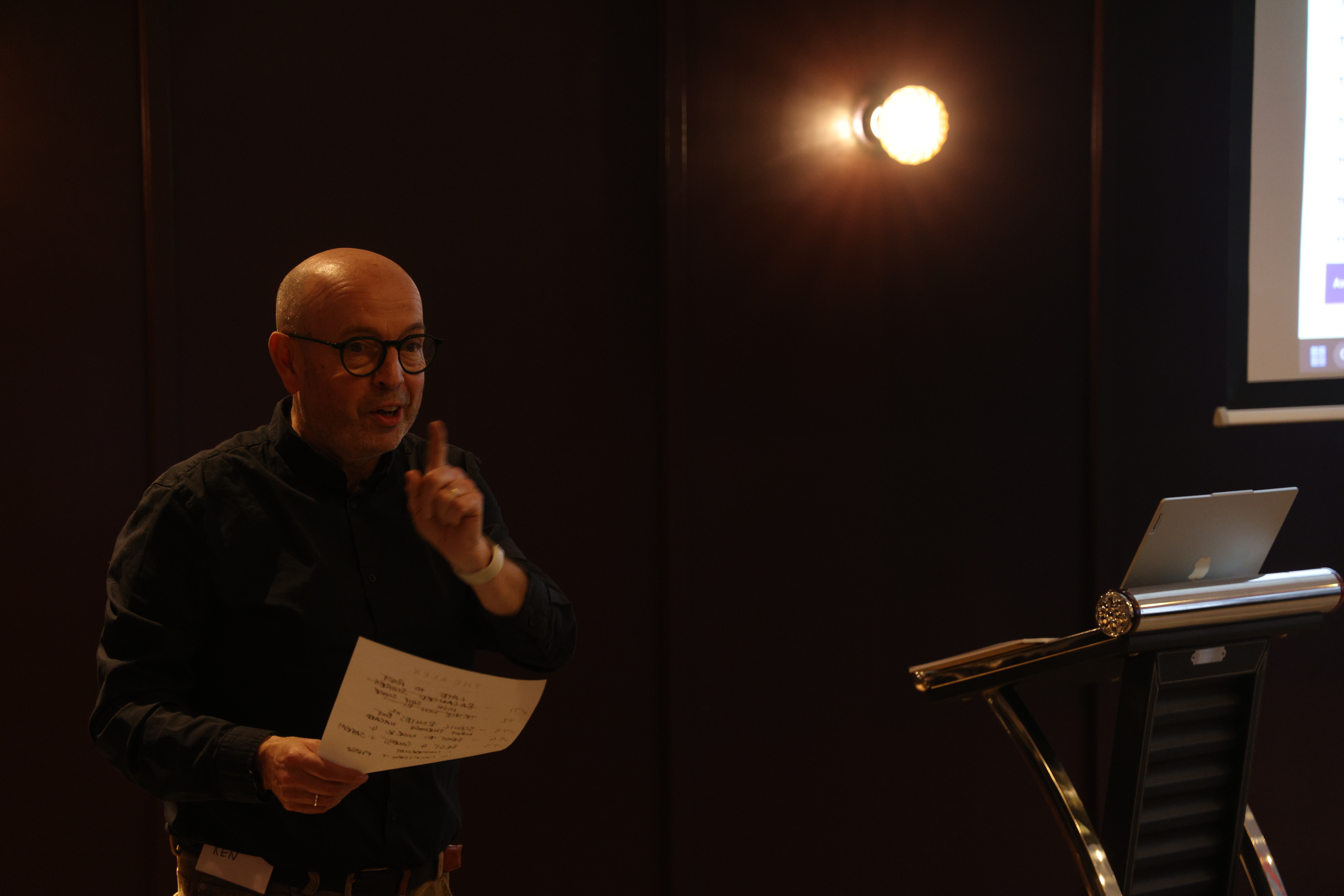 Facilitator presenting to audience during a business simulation, holding notes and gesturing while speaking