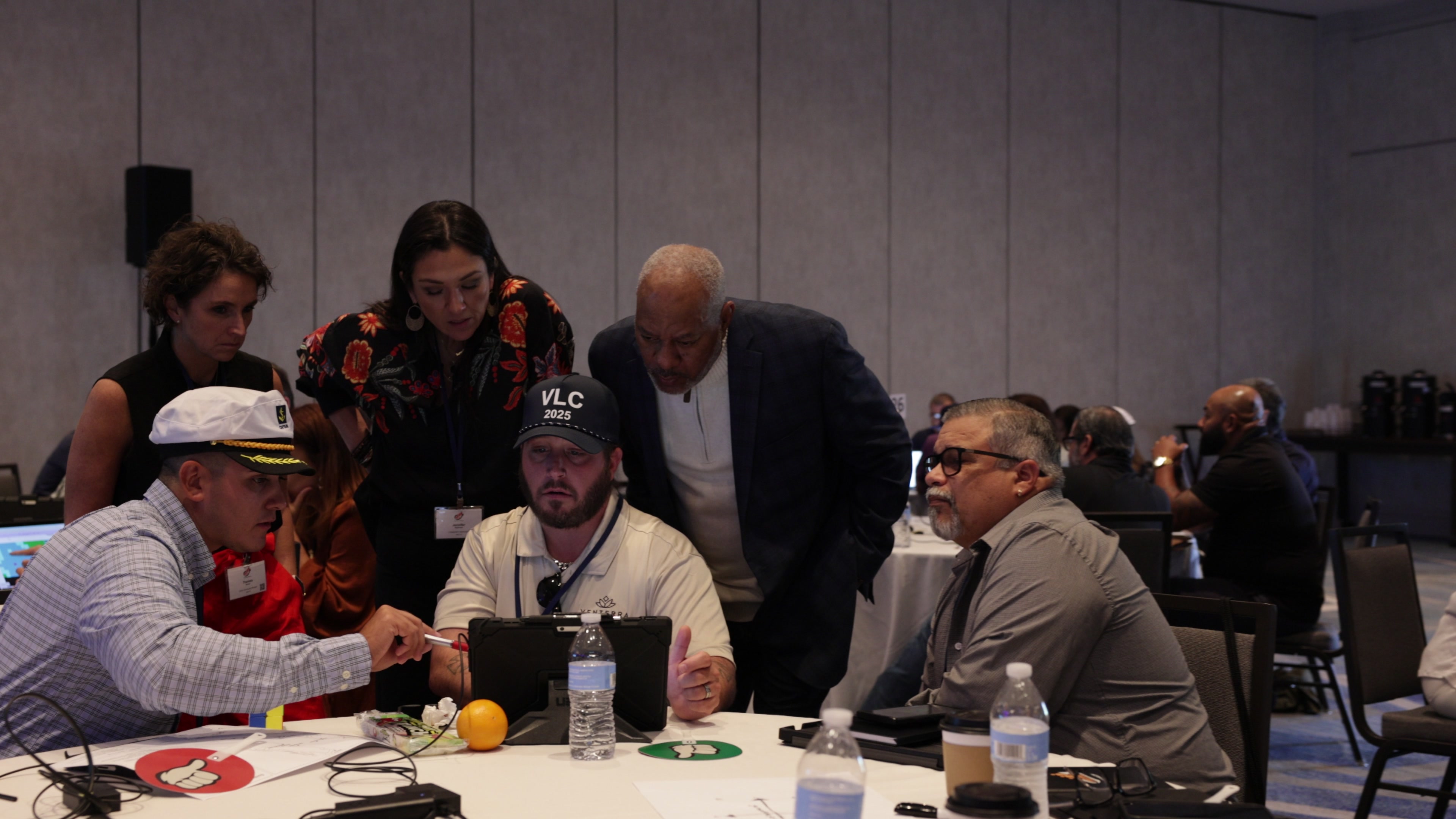 Five people gather around a tablet on a round table, with one person pointing at the screen with a pen.