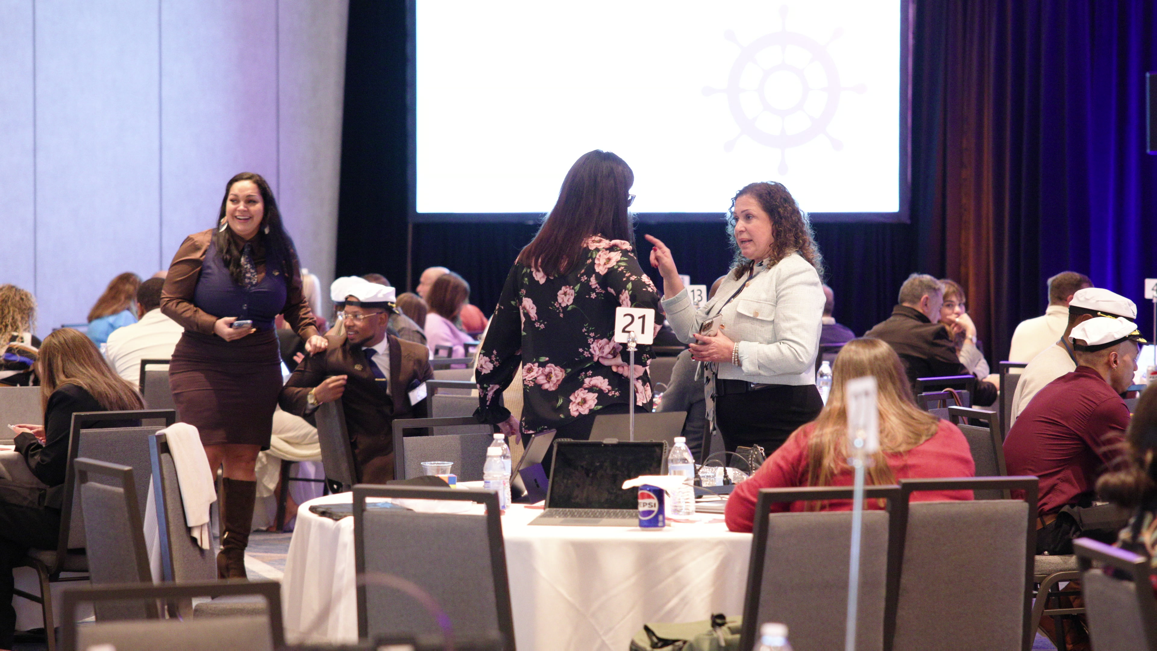 Two women stand and talk in the middle of a conference room while other participants sit at round tables.