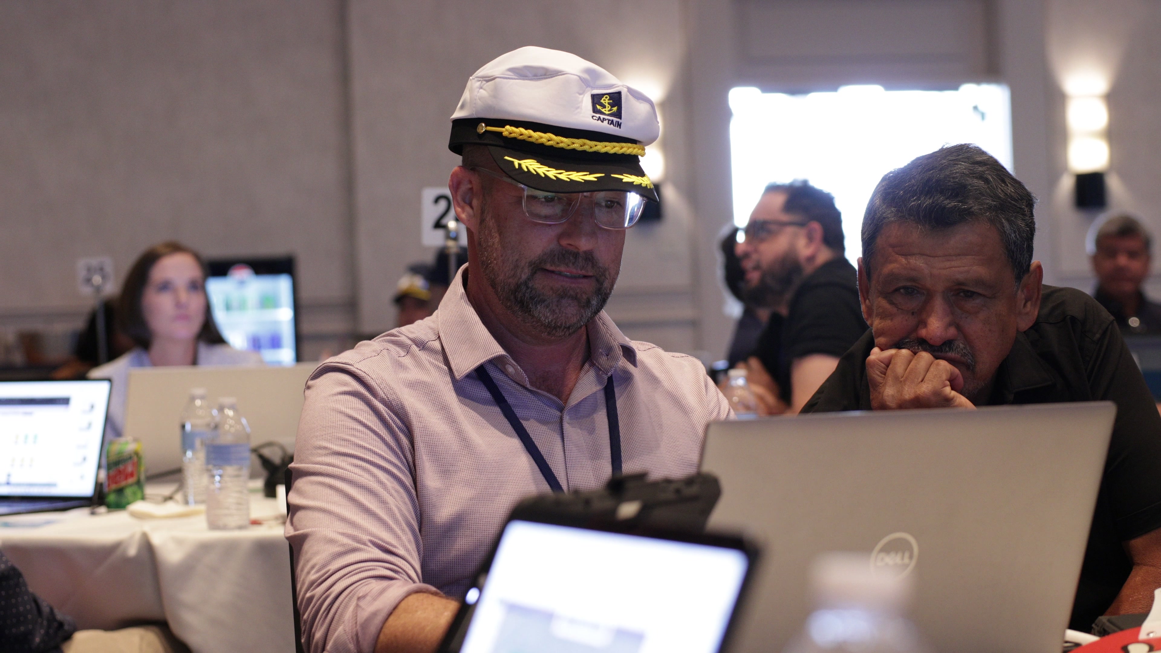 Two men sit at a table looking intently at a laptop screen, with the man on the left wearing a white captain's hat.