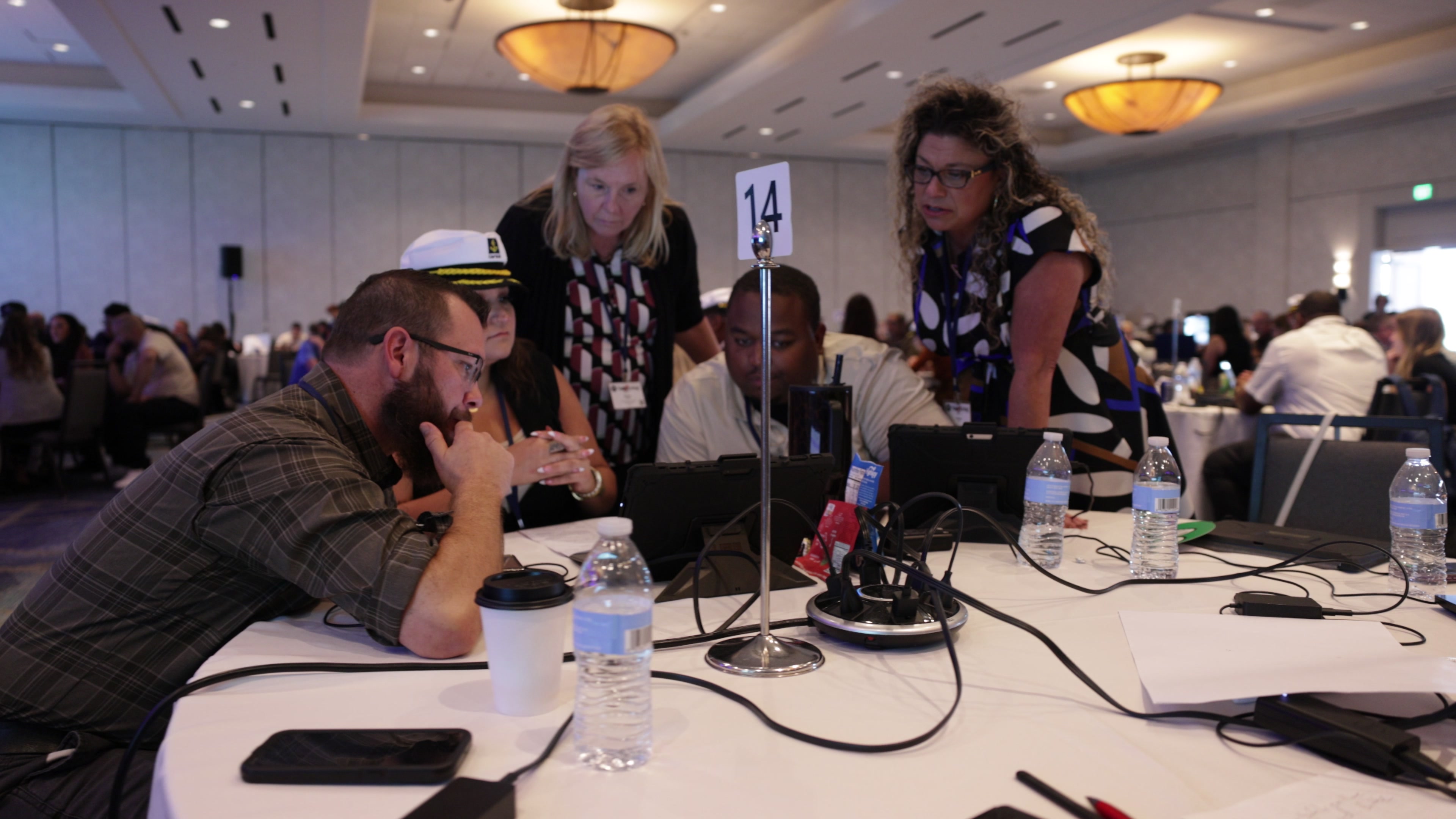 Four people gather around a tablet on a round table in a conference room, looking intently at the screen.
