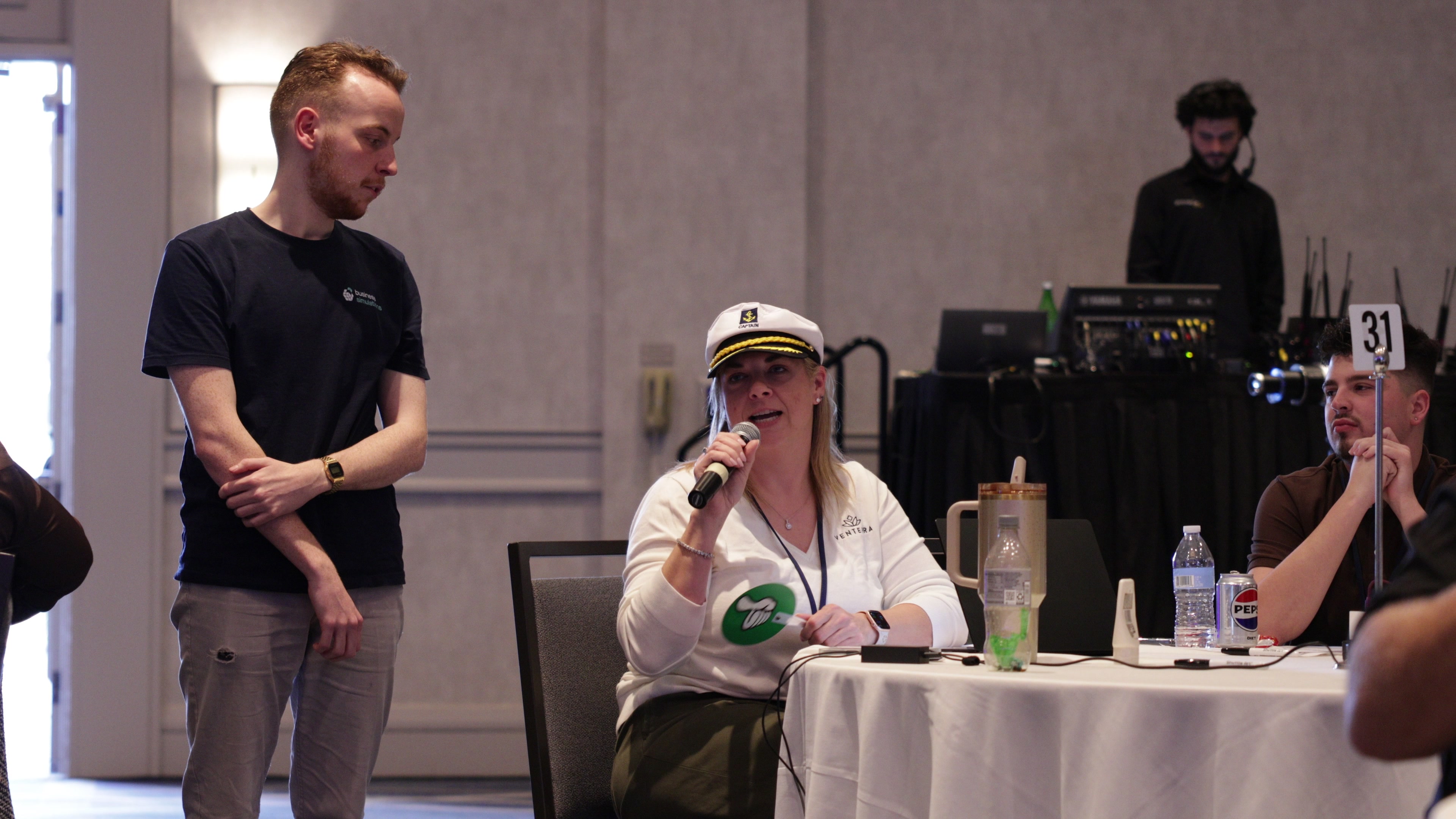A woman wearing a captain's hat speaks into a microphone while seated at a round table, as a man standing next to her listens.