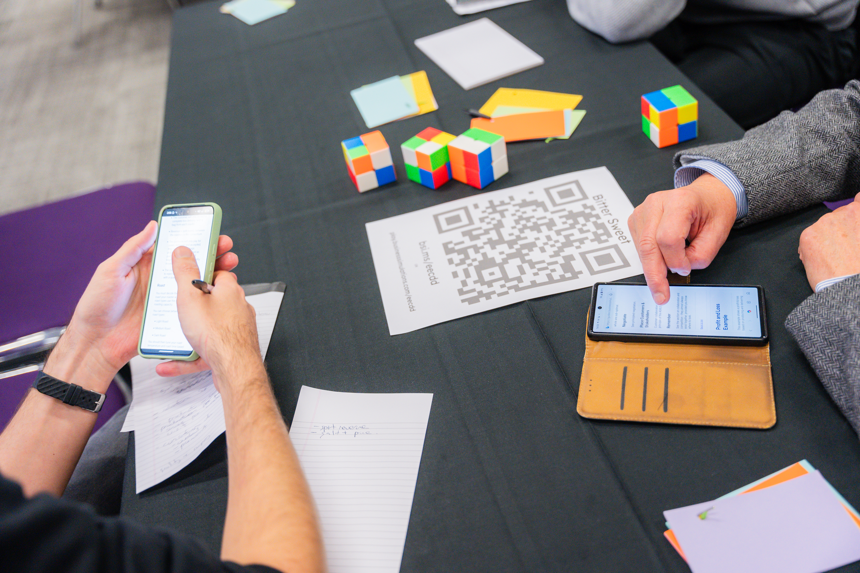 Team members using smartphones and analyzing puzzle cubes and QR codes on a dark table during a business simulation activity.