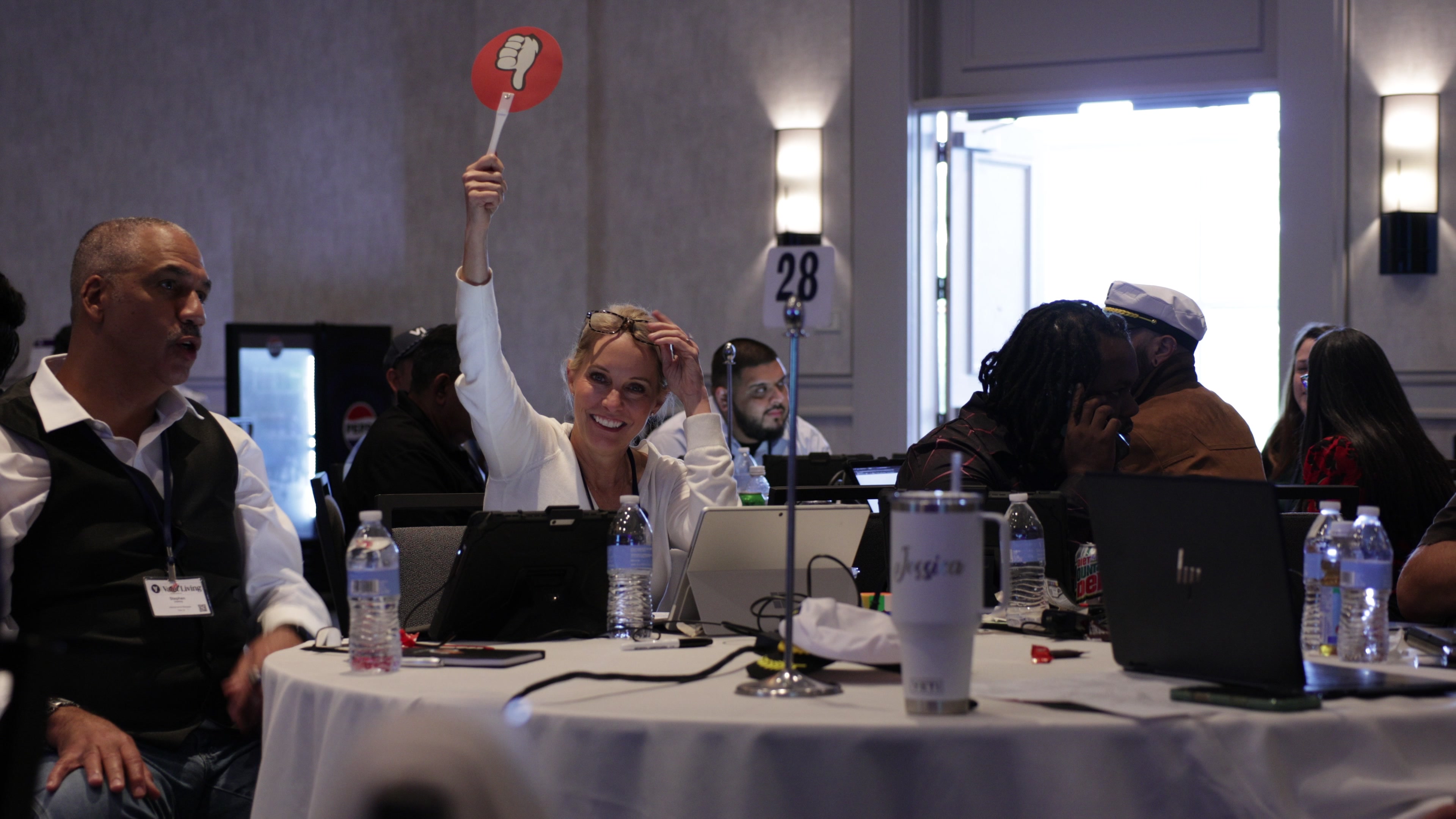 A woman at a round table in a conference room smiles while holding up a red paddle with a thumbs down symbol.