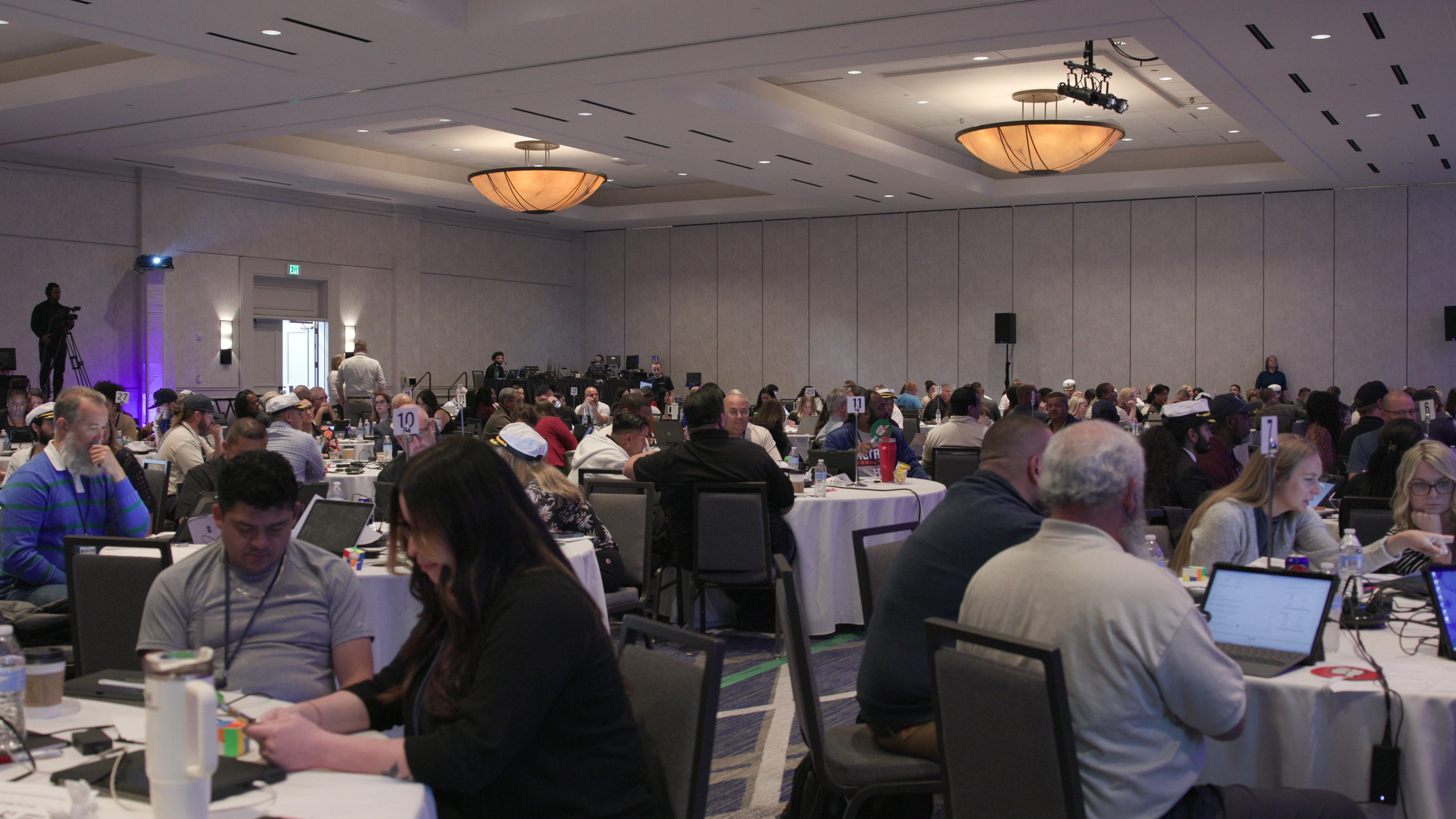 A wide view of a large conference ballroom filled with people sitting at round tables working on laptops and tablets.