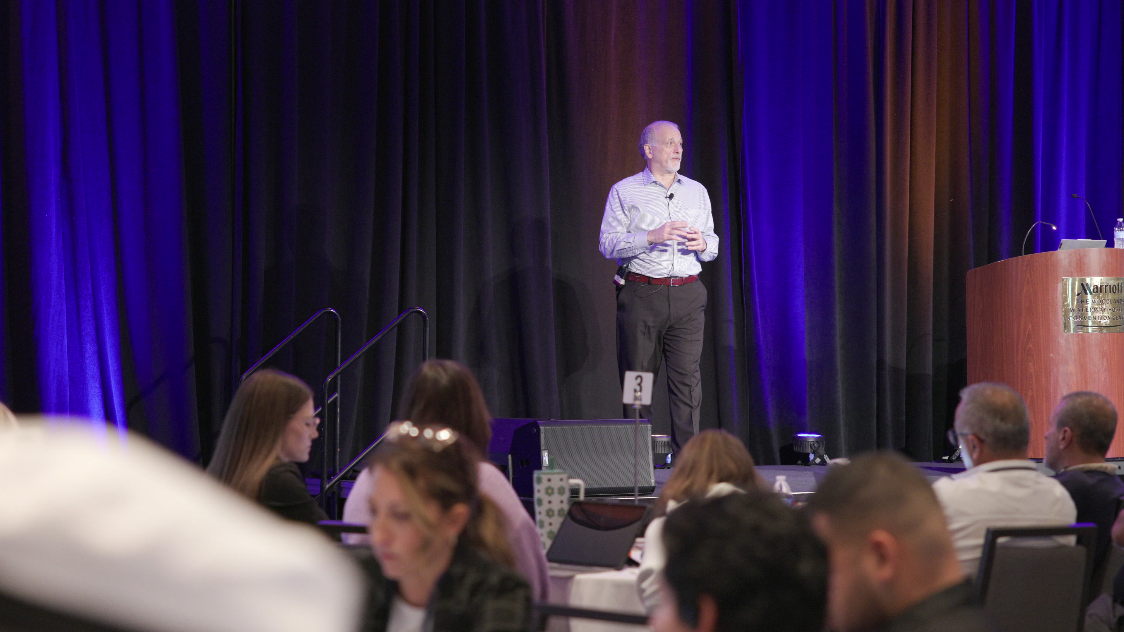 A man stands on a stage presenting to an audience seated at tables in a conference ballroom.