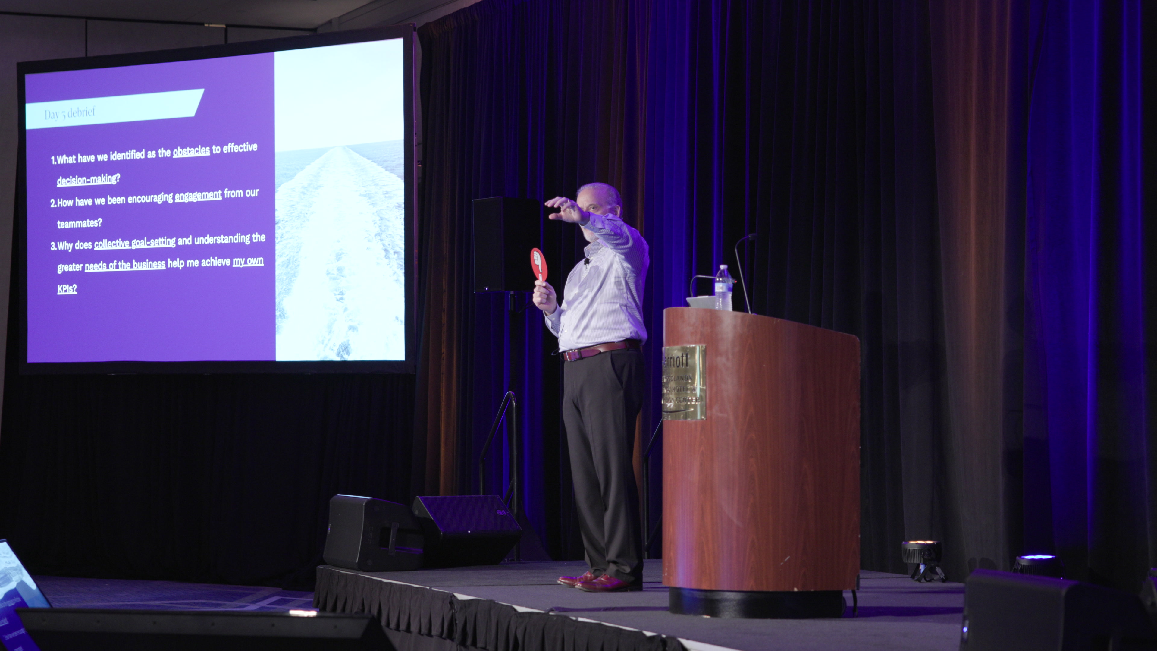 A man stands on a stage next to a wooden podium, gesturing while speaking in front of a large projector screen displaying a presentation.