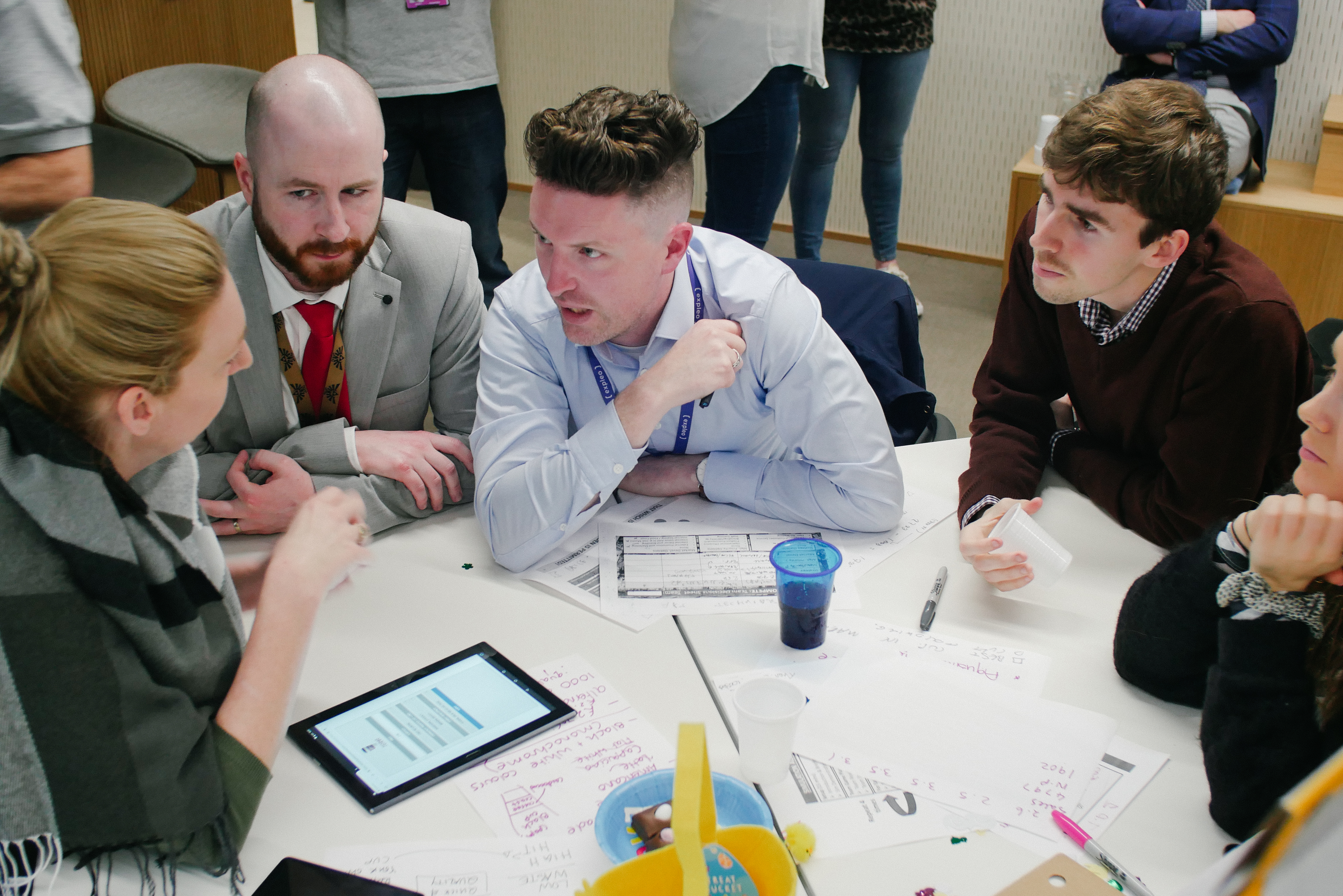 Team members engaged in discussion around a table during a business simulation exercise with documents and tablet.