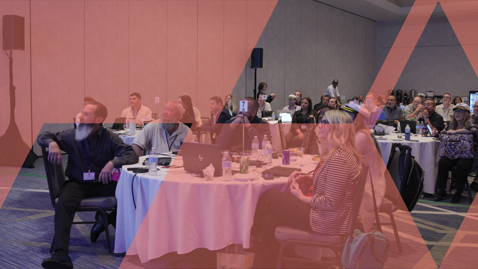 Participants seated at round tables across a conference room, listening during a business simulation session.