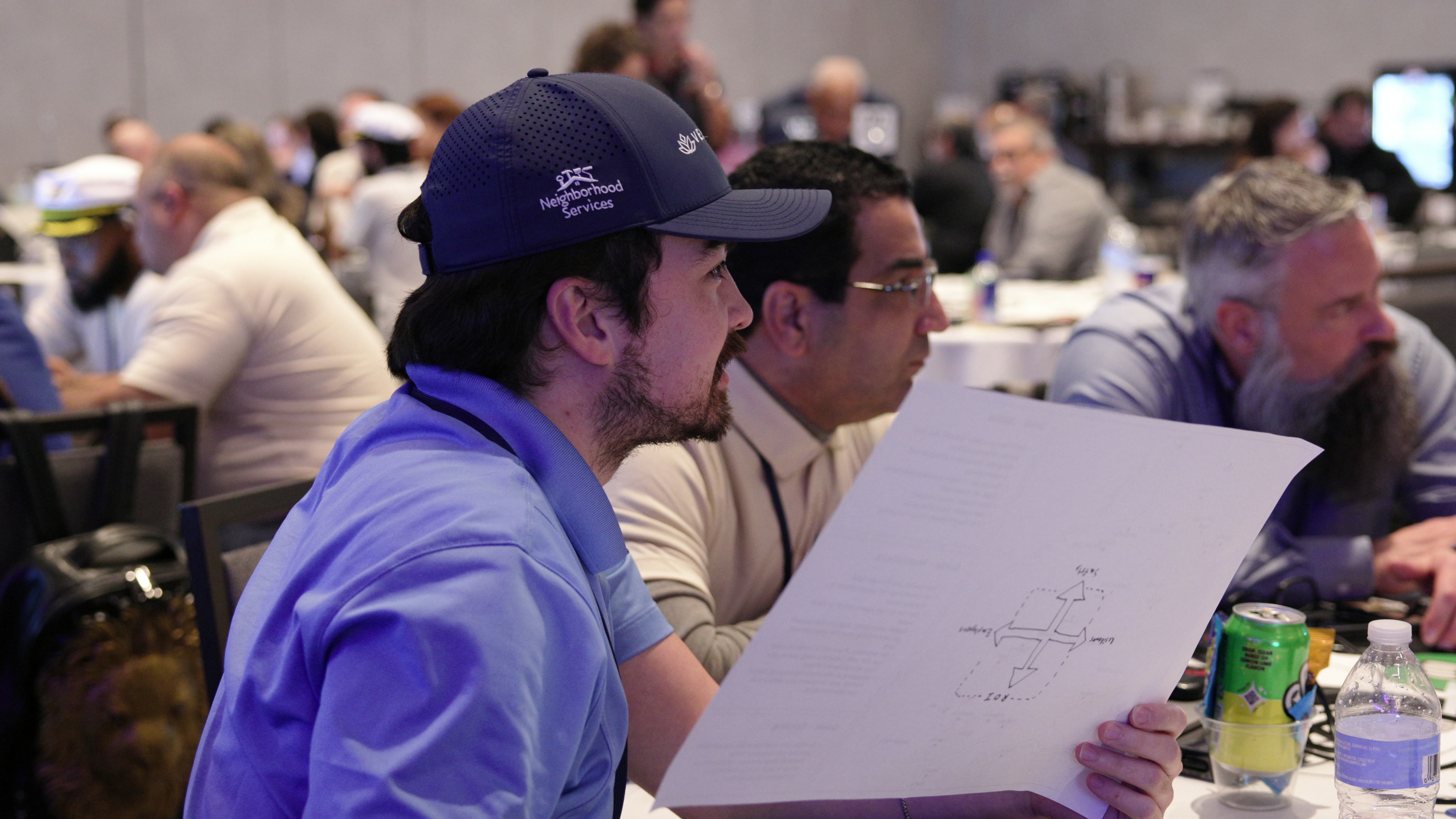 A man in a blue cap holds a piece of paper with a hand-drawn diagram while sitting at a table in a busy conference room.