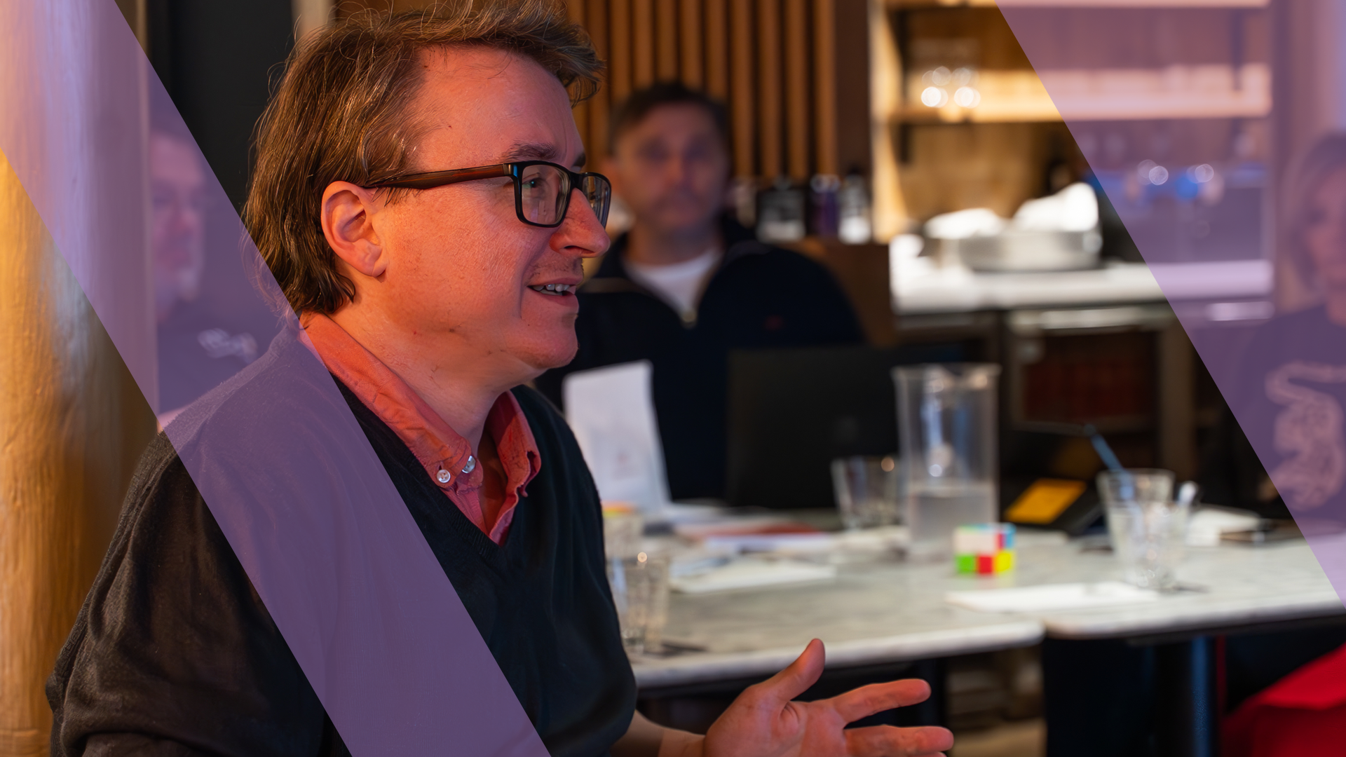 A team member speaking candidly during a business simulation while a colleague listens, overlaid with a purple Business Simulations brand motif