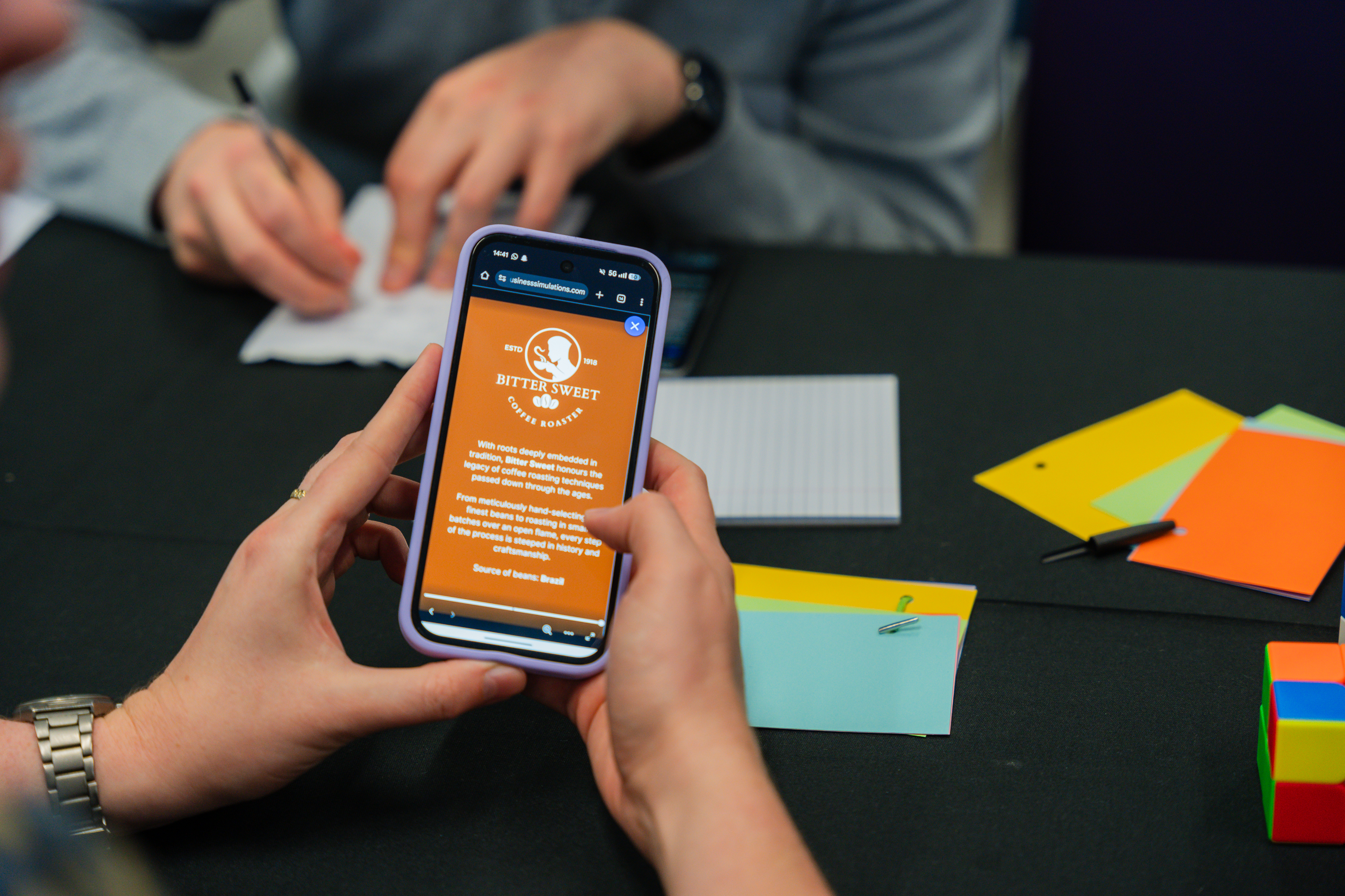 Team members view a mobile app on a smartphone during a business simulation workshop with colorful sticky notes on the table.