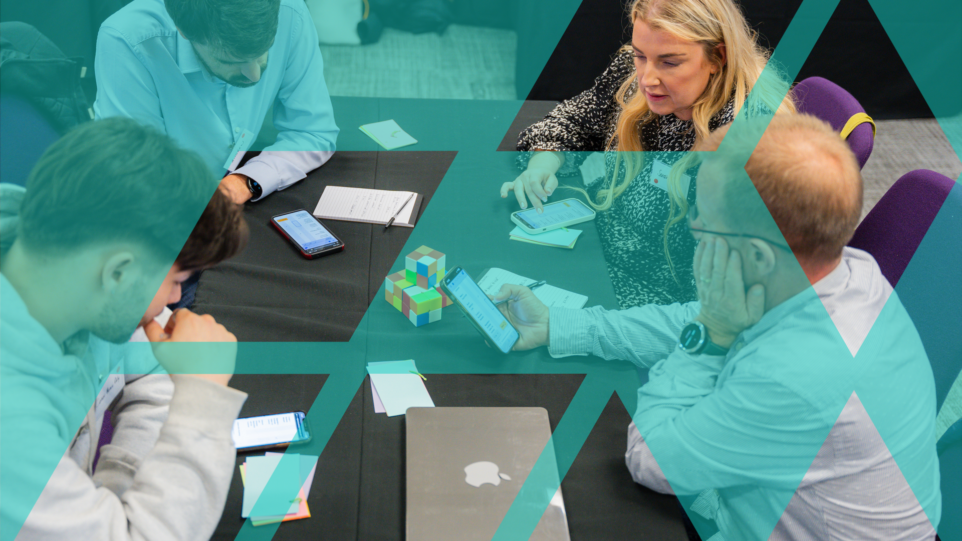 Four participants in a simulation discussing at a table while looking at the simulation on a phone - watermarked with the Business Simulations motif