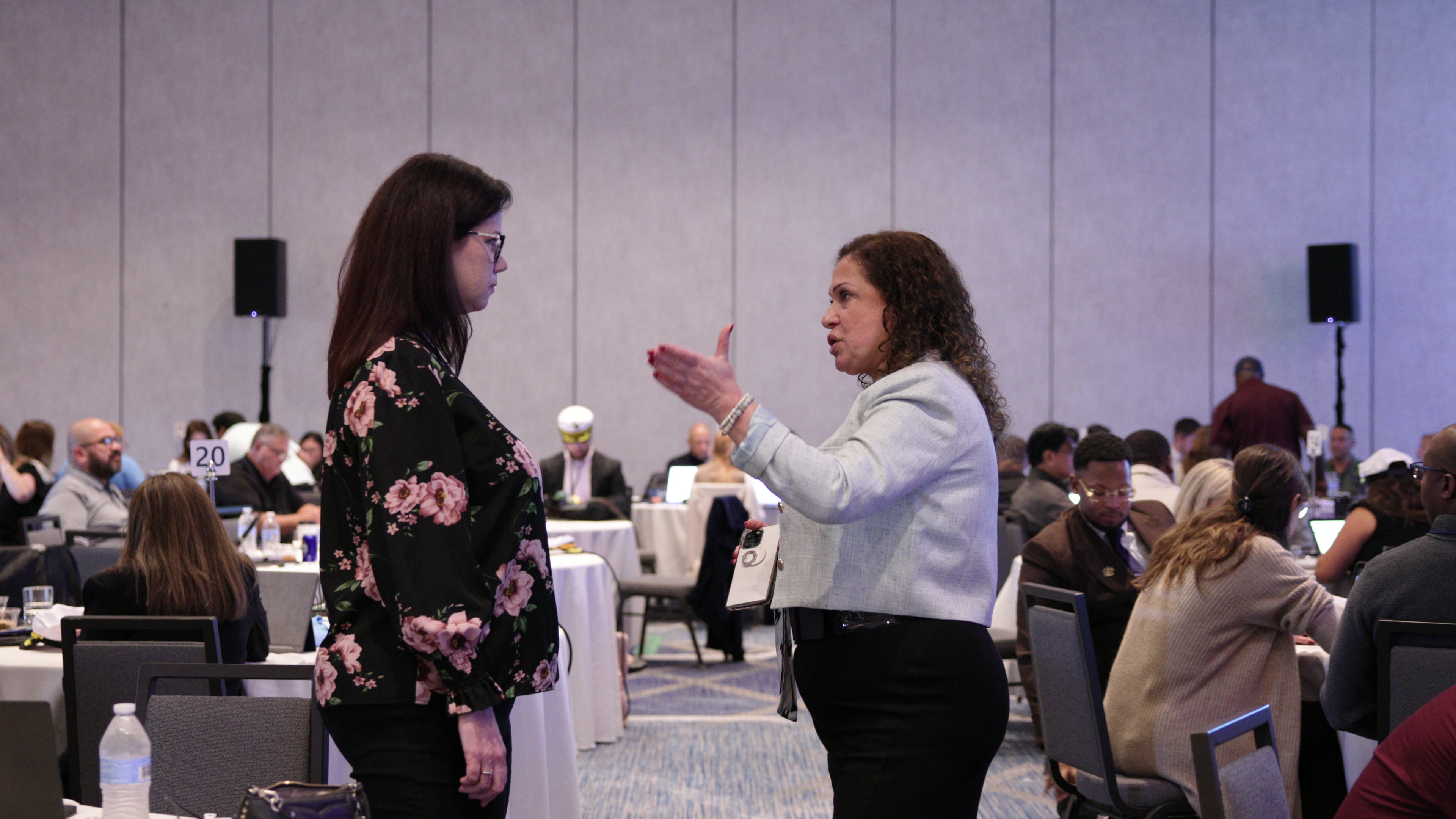 Two women stand facing each other in a conference room, one gesturing with an open hand as she speaks while the other listens.