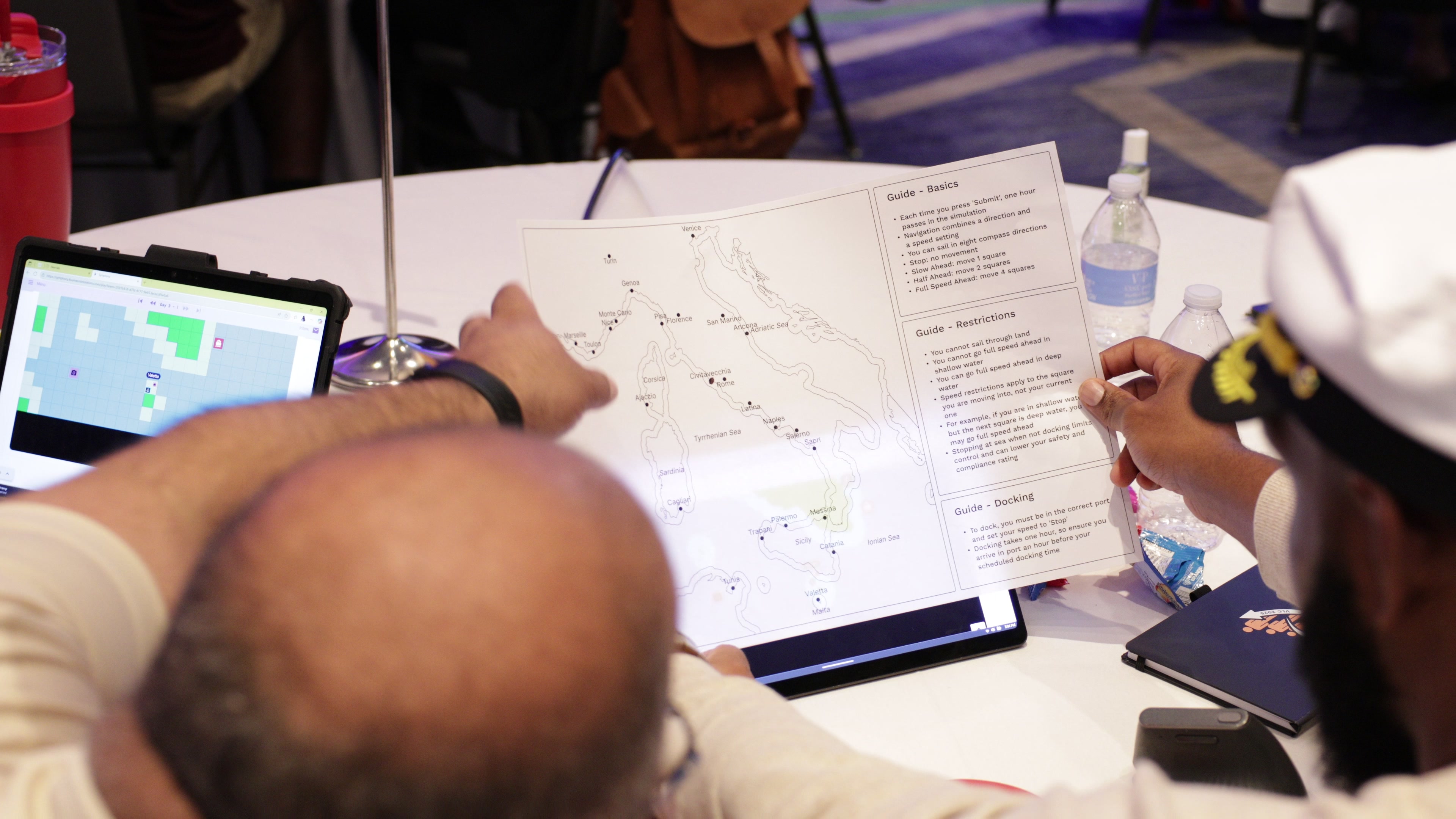 Over-the-shoulder view of two people at a table looking at a map and instructions on a large tablet screen.