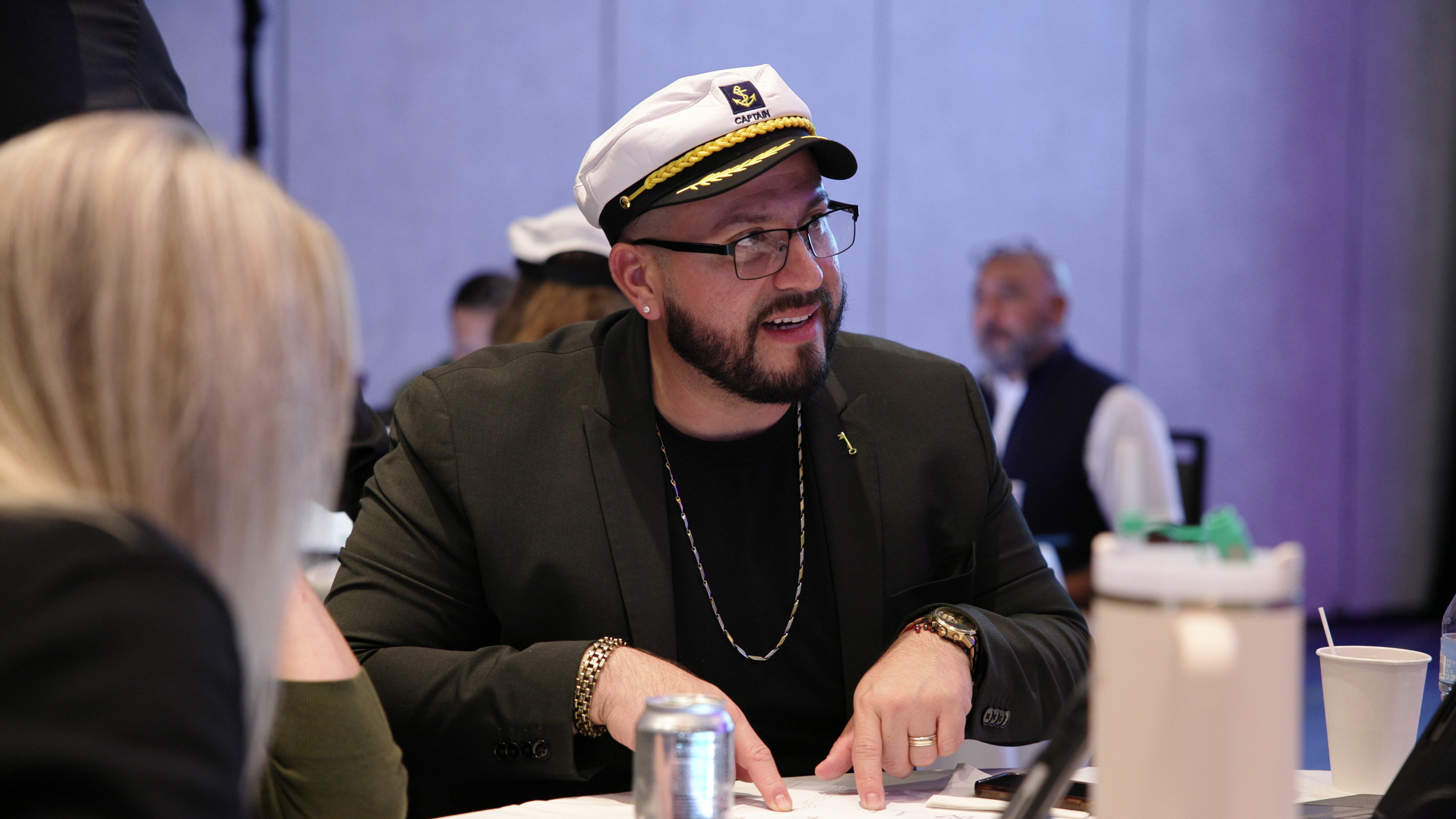 A man wearing a naval captain's hat and glasses sits at a round table, pointing at a document while talking to a colleague.