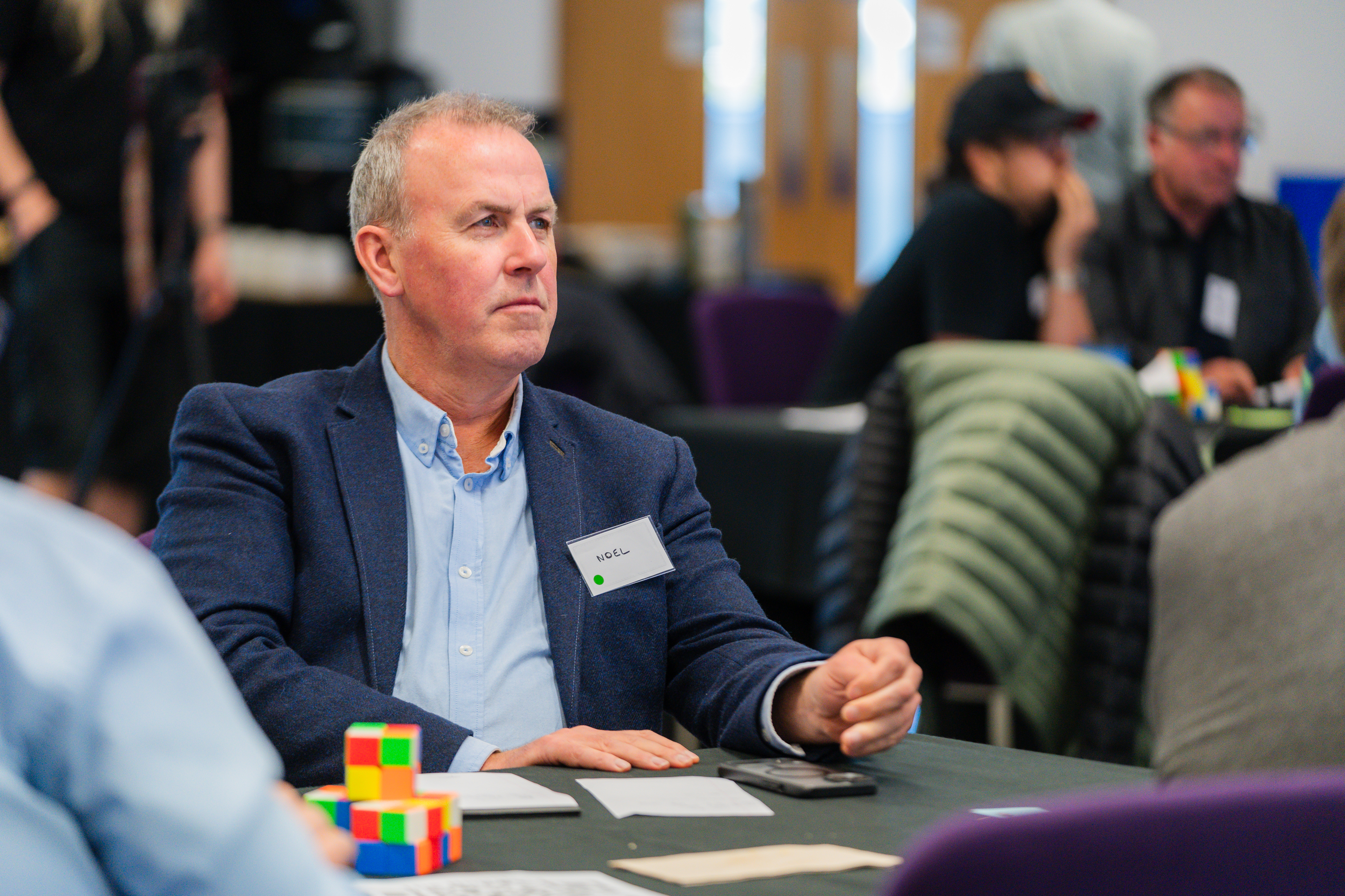 Business simulation participant in navy blazer engaged in strategic discussion at team table with colorful building blocks.