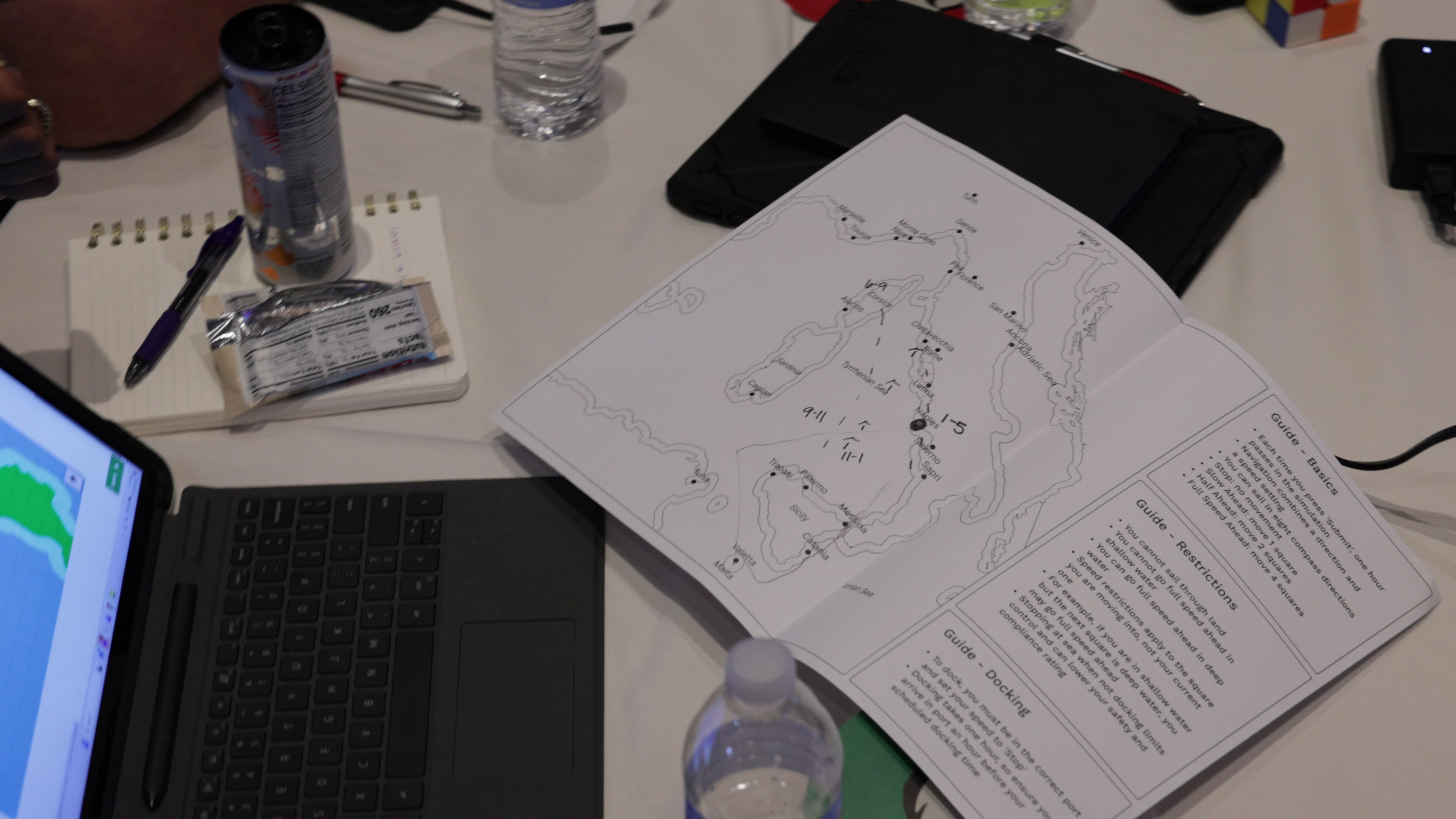 A close-up view of a table showing a printed map with markings, a laptop displaying a digital map, a notebook, and various drinks.