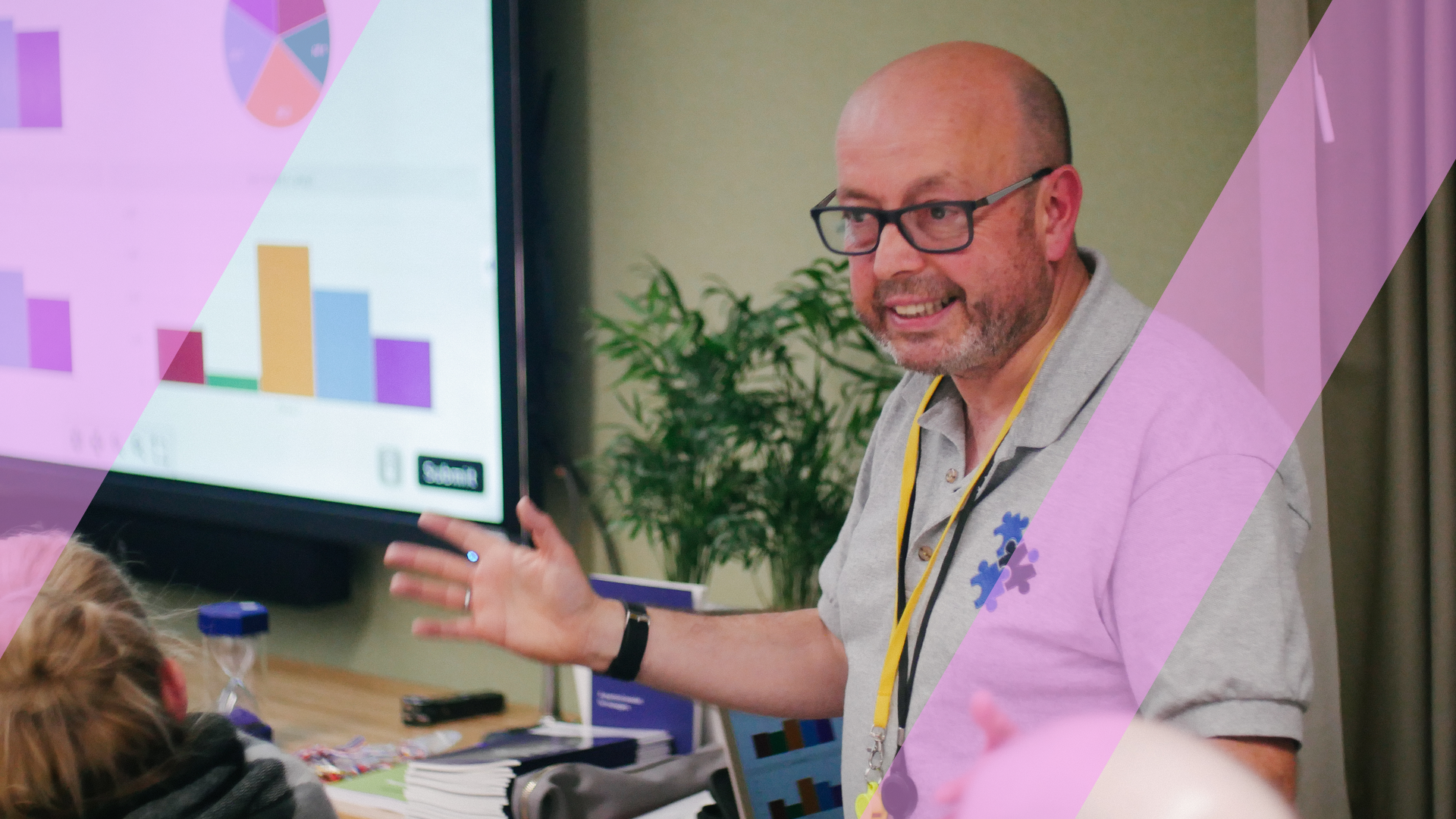 A facilitator gesturing to a live business simulation dashboard on a large screen while presenting to participants, overlaid with a pink Business Simulations brand motif