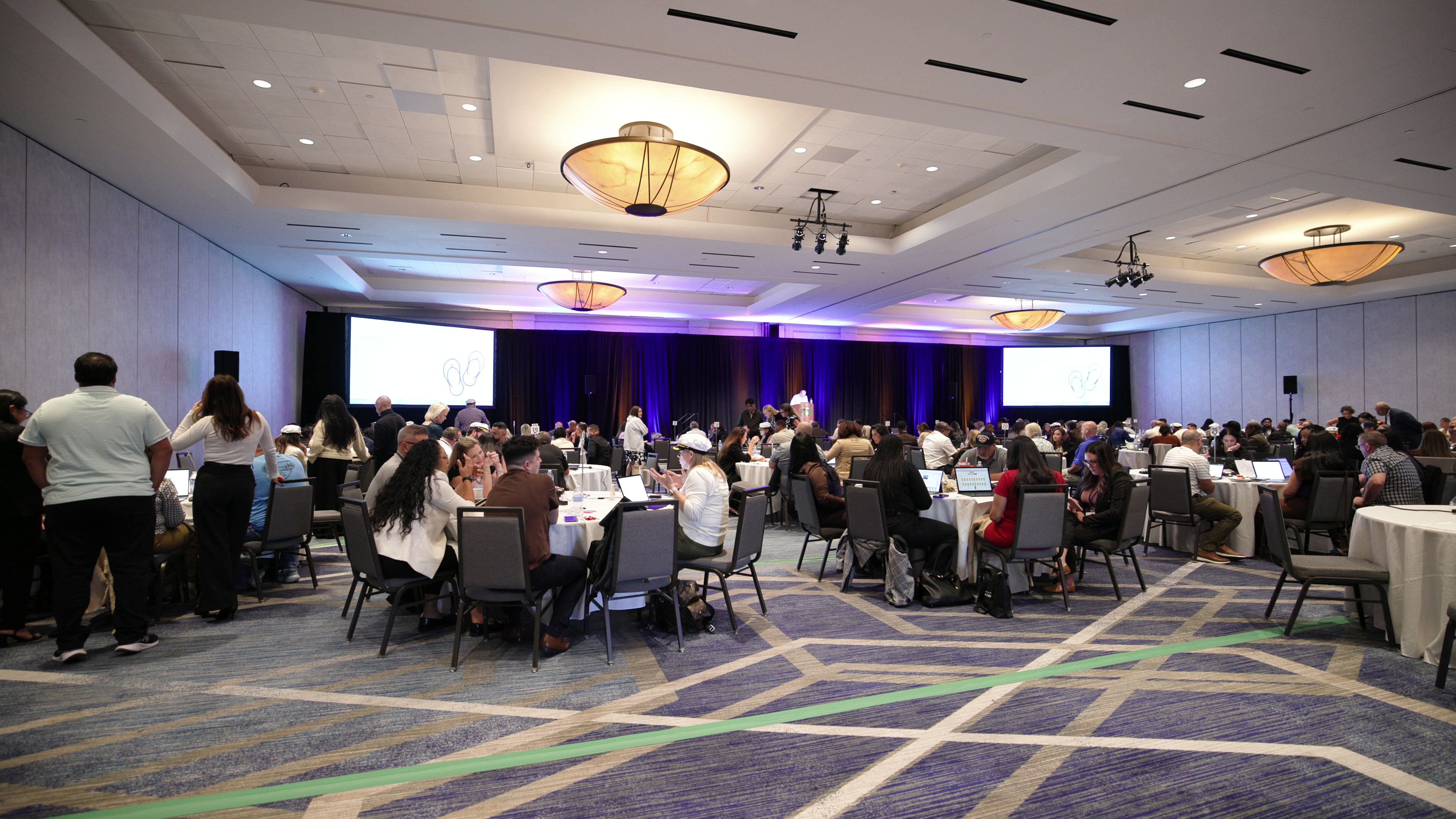 A wide view of a large conference ballroom filled with people sitting at round tables, using laptops and discussing.