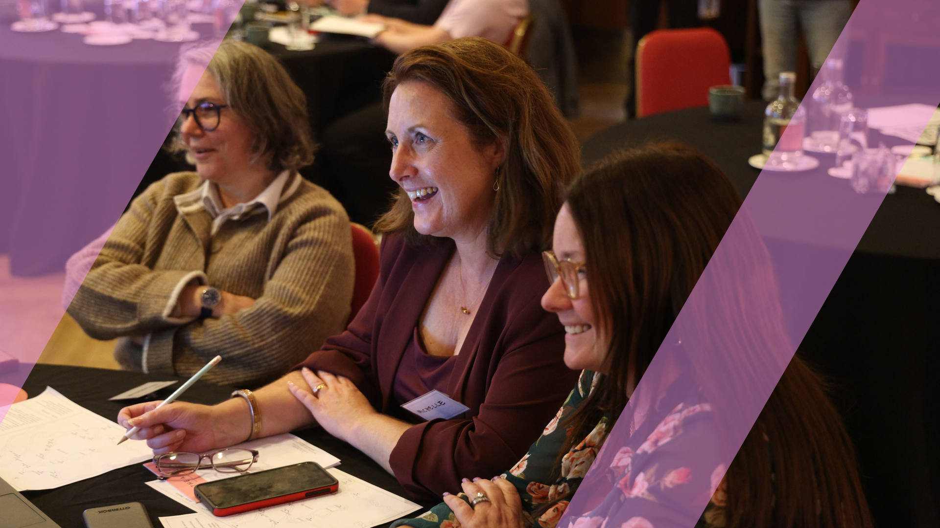 Three participants smiling and engaged at a table during a live, facilitated business simulation, overlaid with a pink Business Simulations brand motif