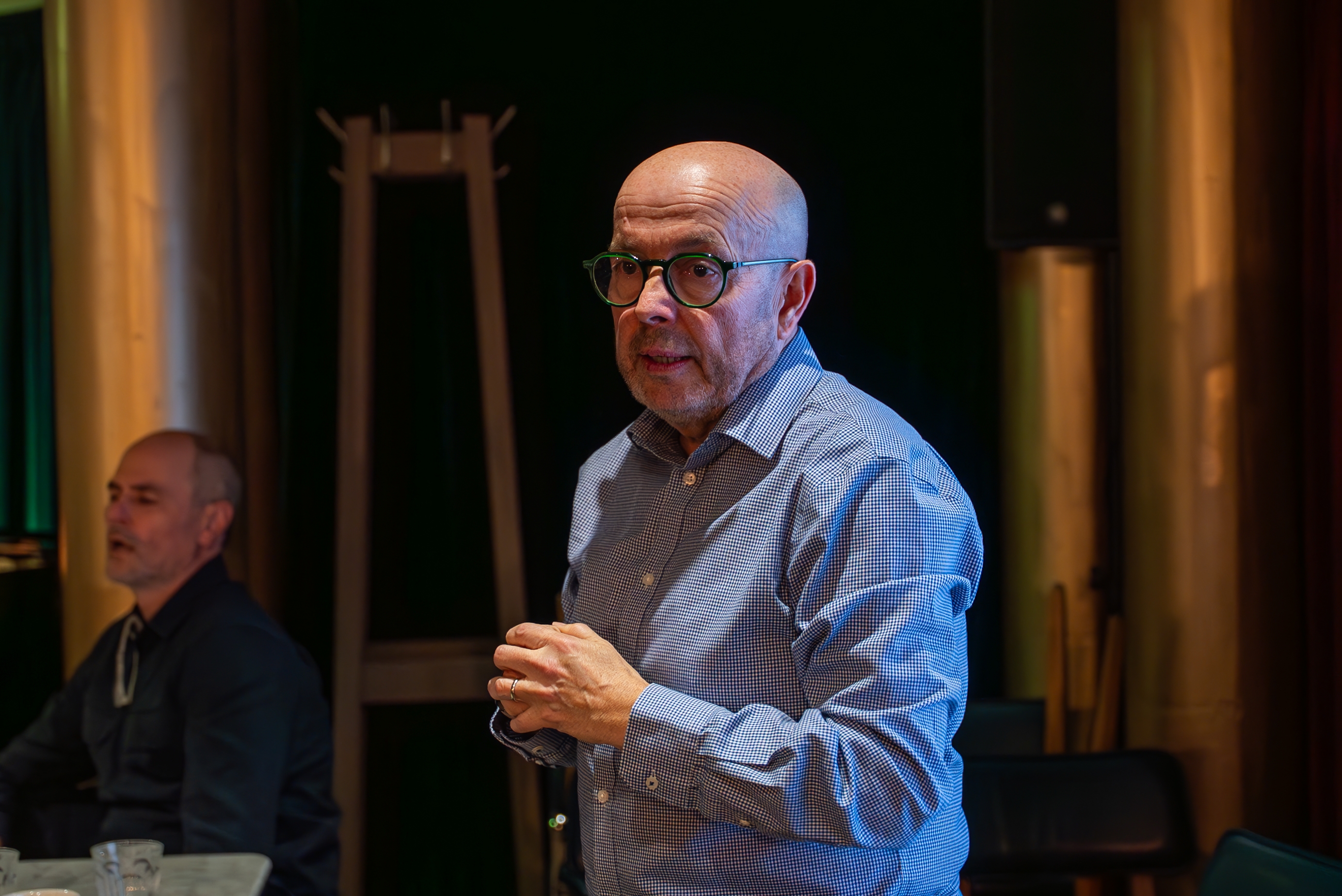 Facilitator in blue shirt and glasses gestures while speaking to business simulation participants.