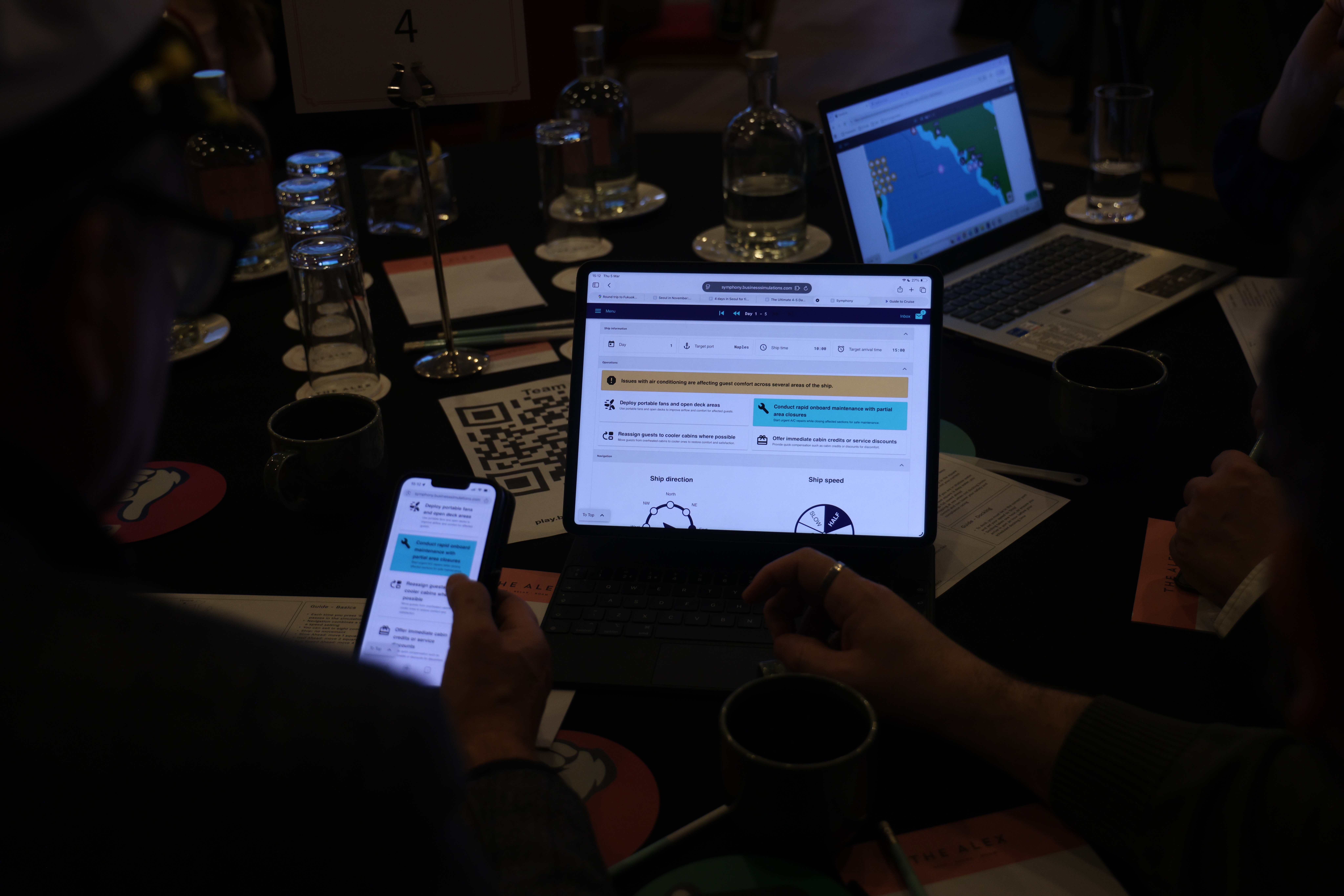 Team members gathered around a table view simulation data on tablets and laptops during a business simulation workshop.