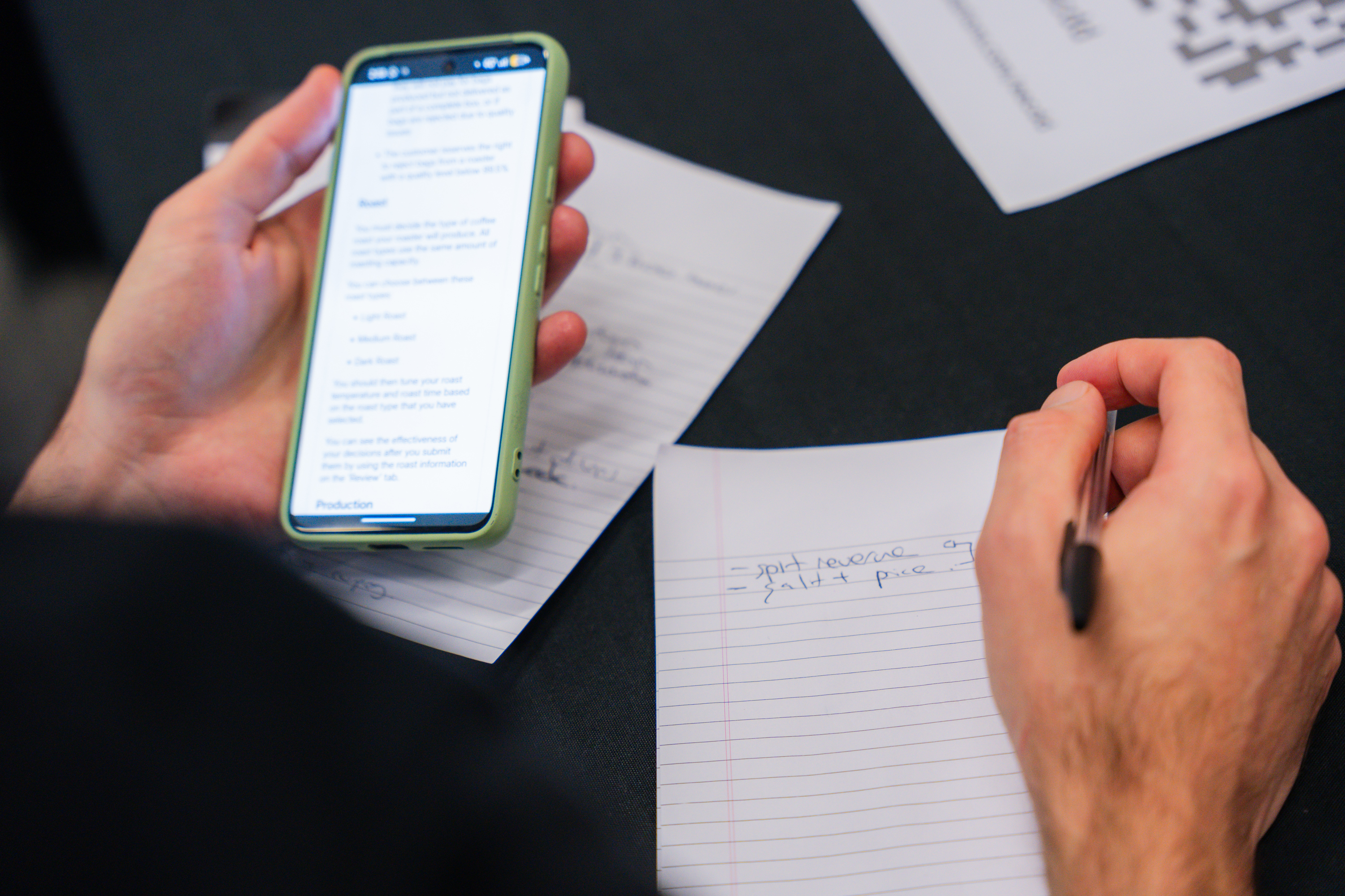 Participant holding a smartphone displaying text while writing notes on paper during a business simulation exercise.