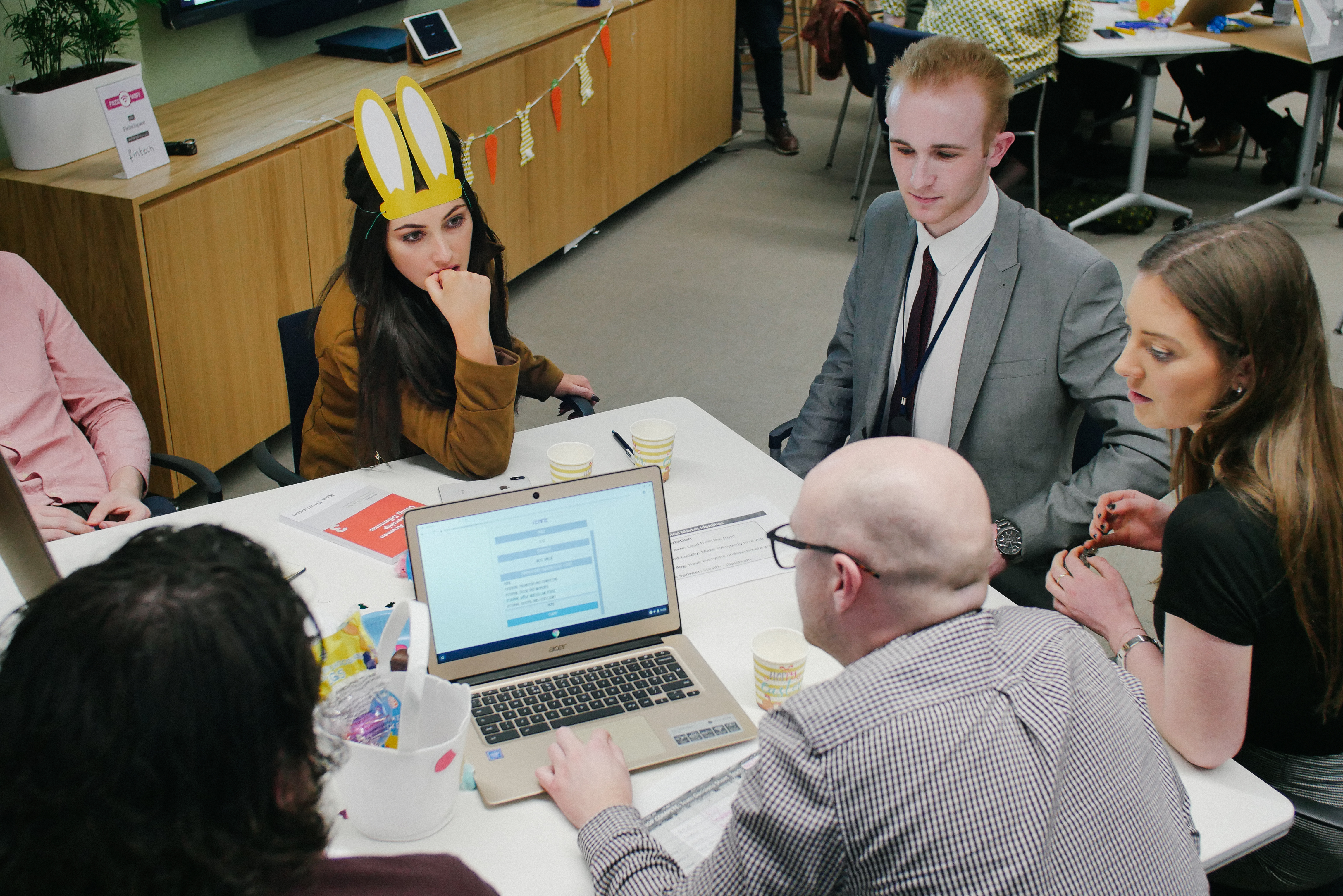 Business simulation team gathered around laptop engaging in collaborative discussion and decision-making.