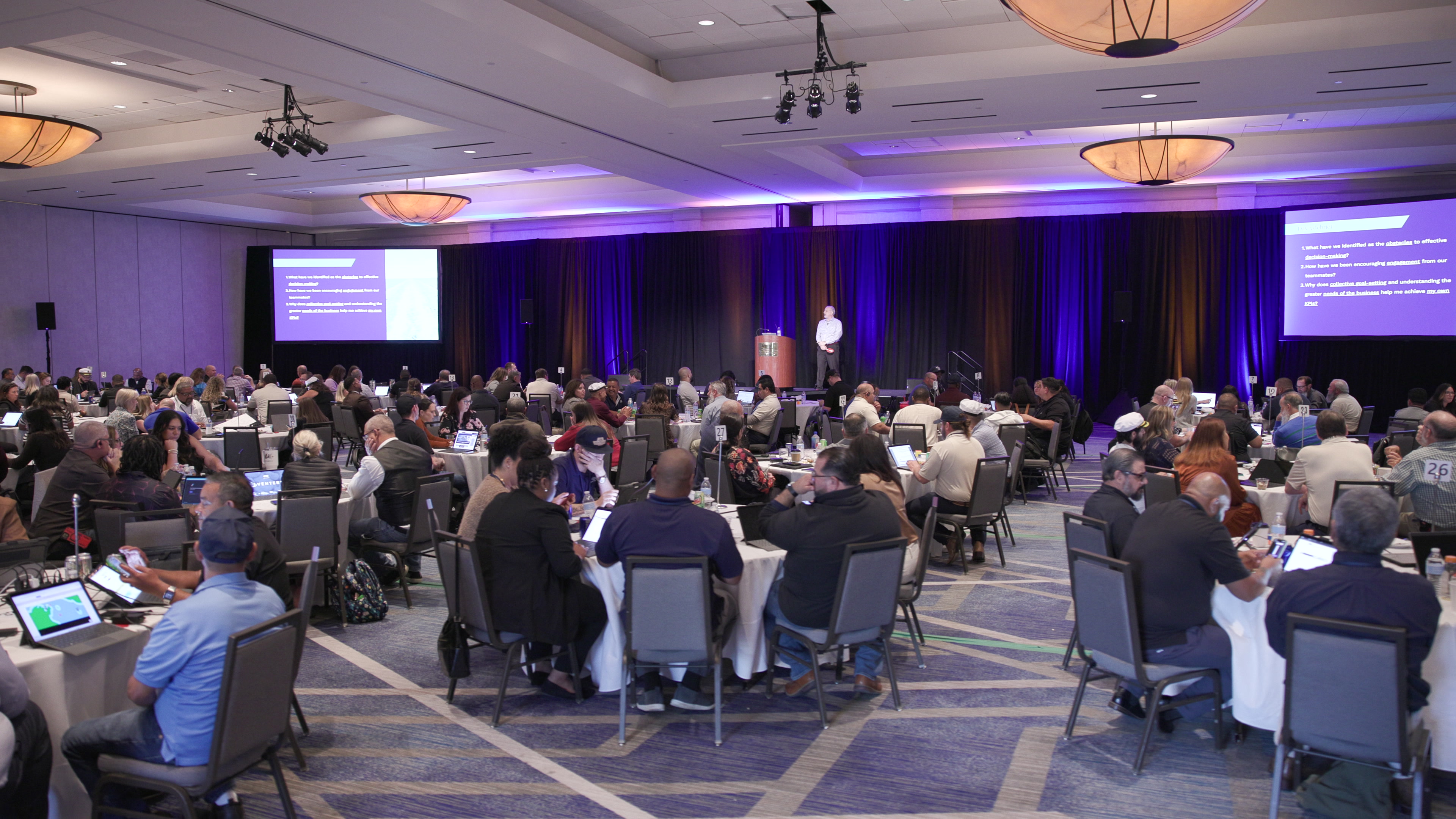 A wide view of a large conference room filled with round tables where participants sit, while a facilitator speaks from a stage with two large projector screens.