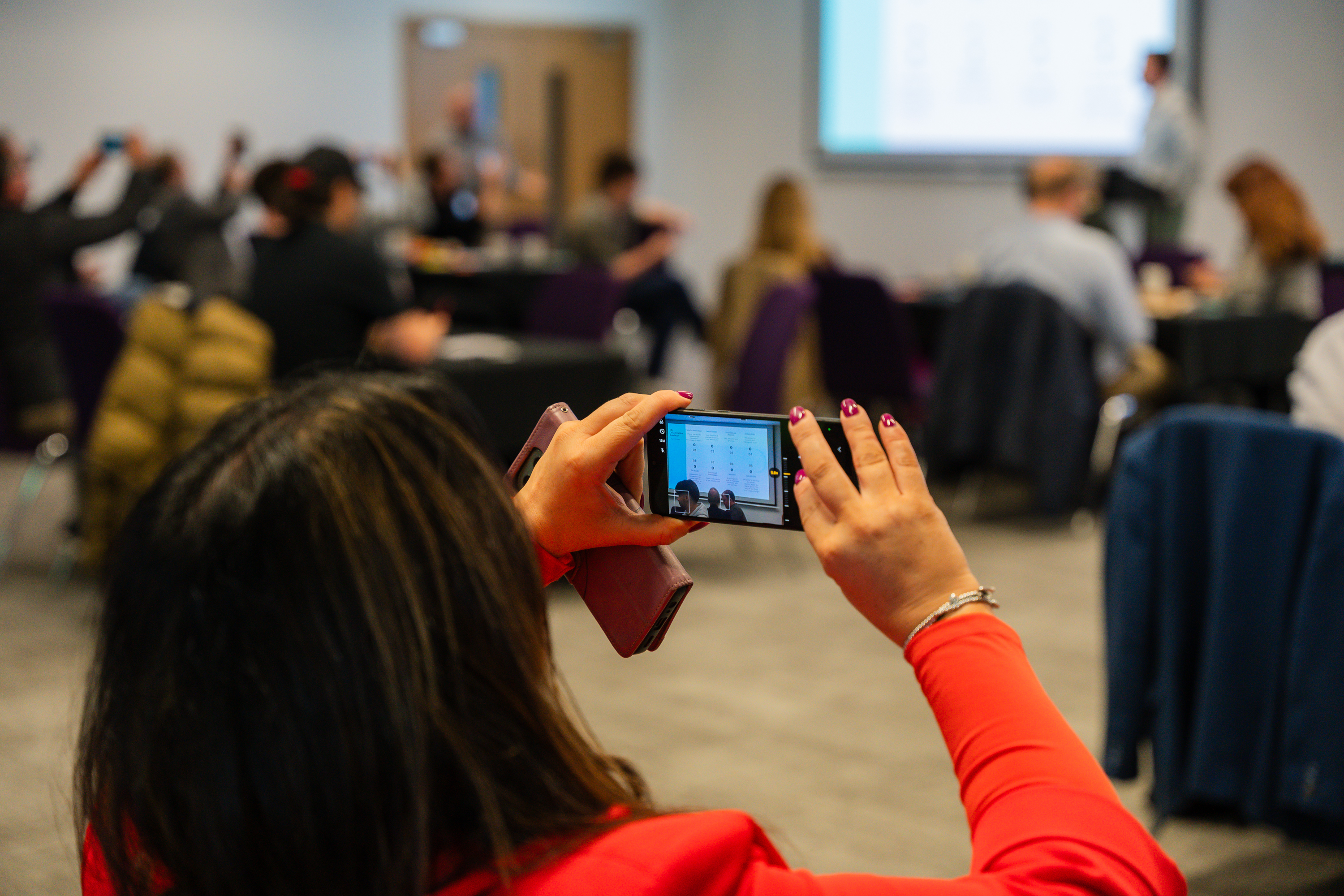 Participant in red jacket photographs a business simulation session with smartphone while other team members engage in background.